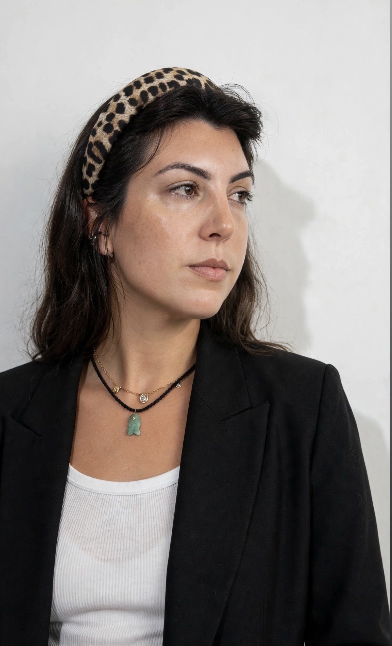 Woman with dark hair wearing a leopard print headband, black blazer, and white top, looking to the side against a plain background.