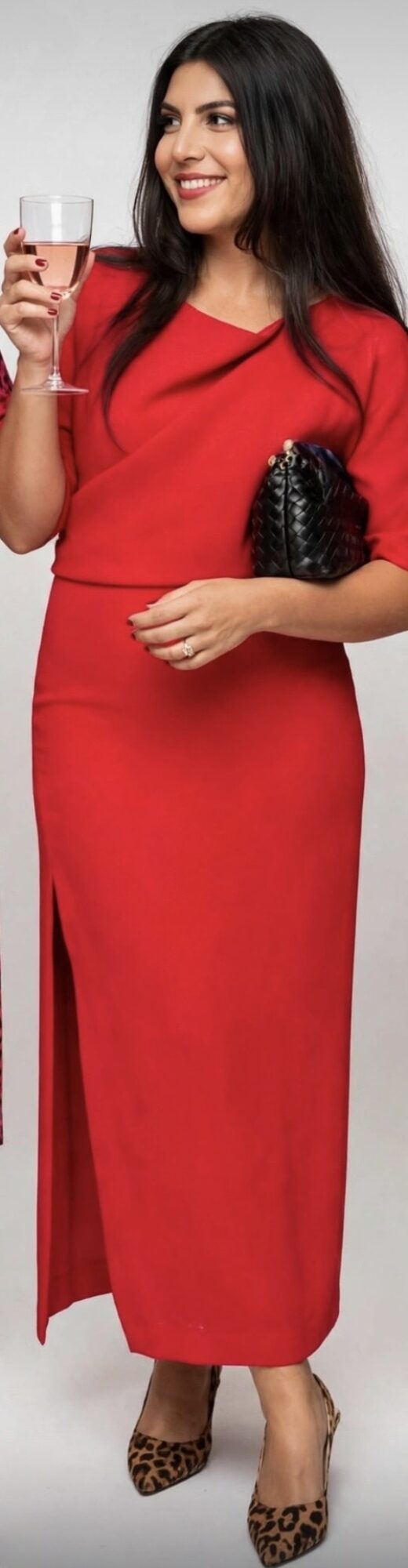 Woman in red dress holding a glass of wine, smiling, with a black clutch, standing against a plain background.