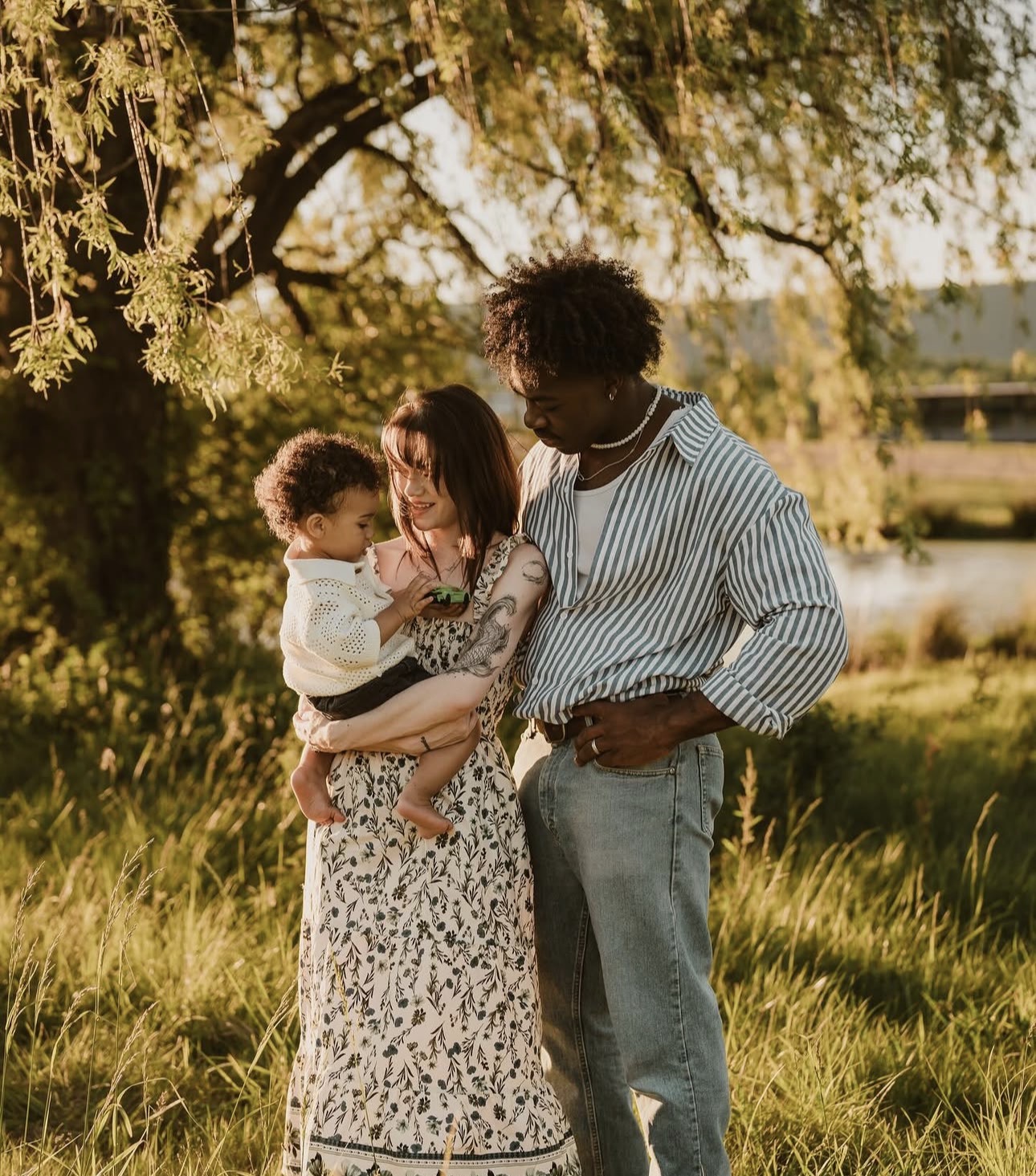 Family of three outdoors under a tree, woman holding a child, man standing beside them, sunlight filtering through leaves.