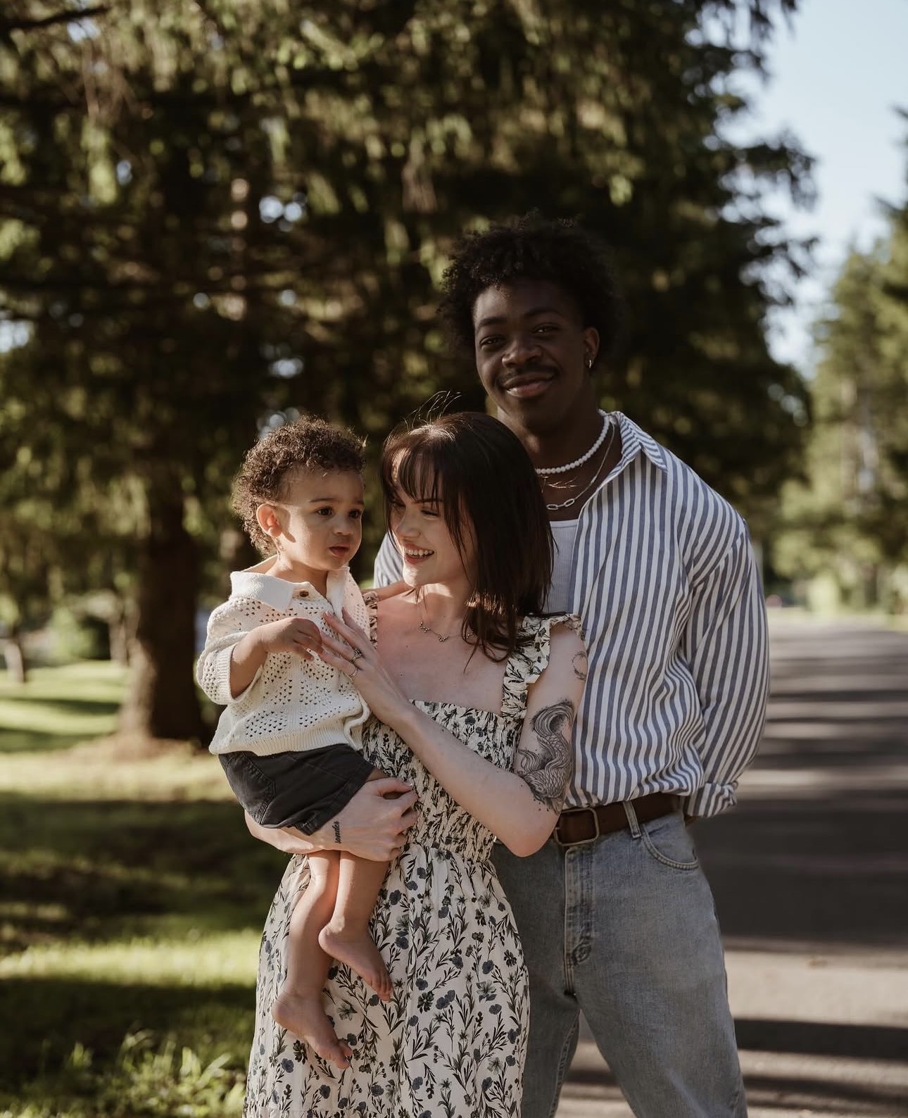 Three people outdoors, a woman holding a child, and a man standing beside them, trees in background, sunny day.