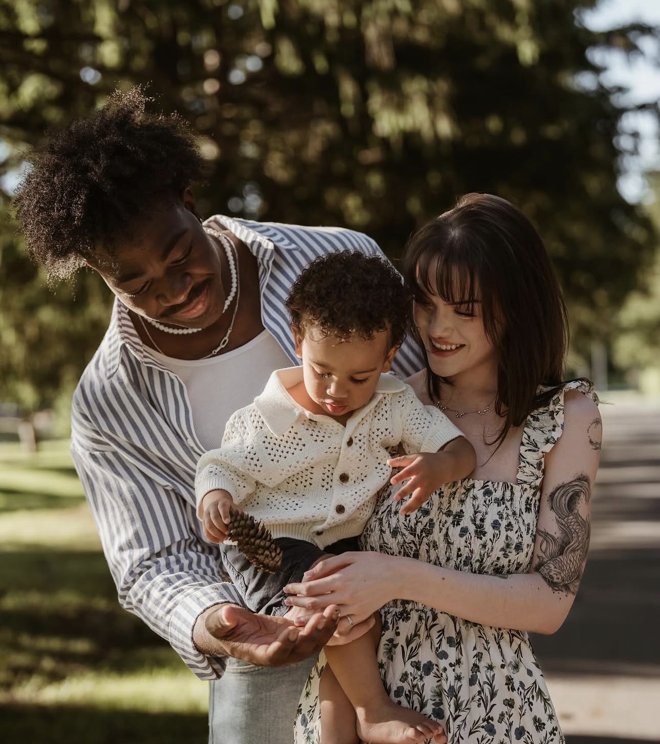 Three people, a man, a woman, and a child, smiling and looking at a leaf outdoors with trees in the background.