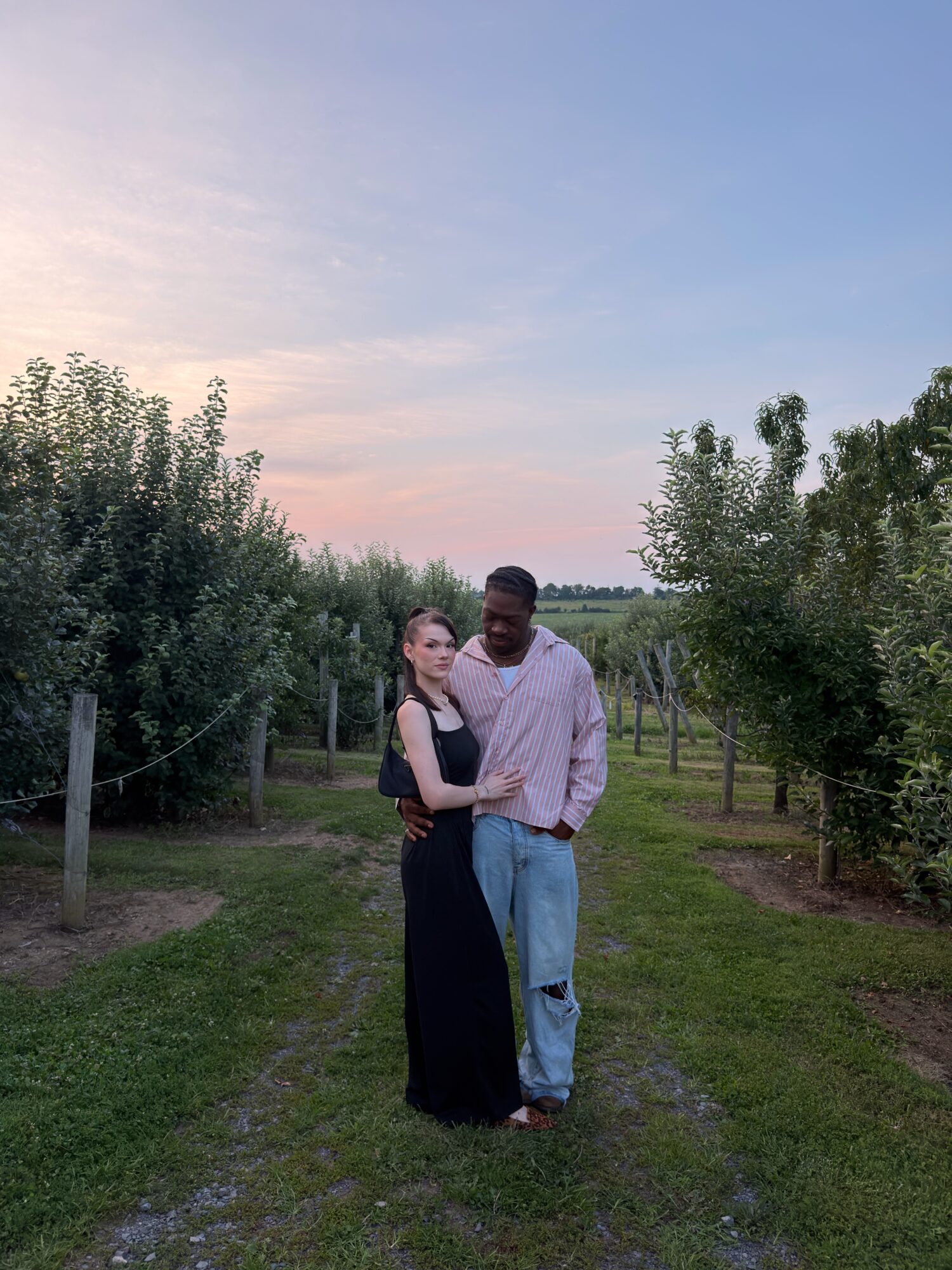 Two people standing outdoors on a grassy path with trees and a sunset sky in the background.