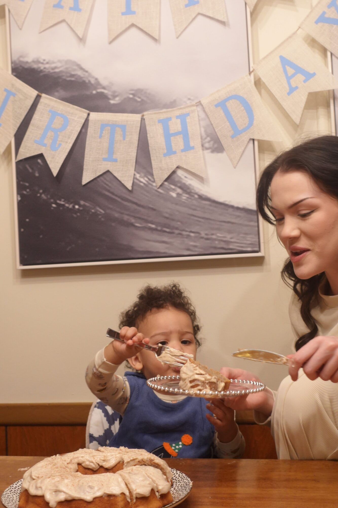 Two children eating cake at a birthday celebration, with a large cake on the table and a mountain picture in the background.
