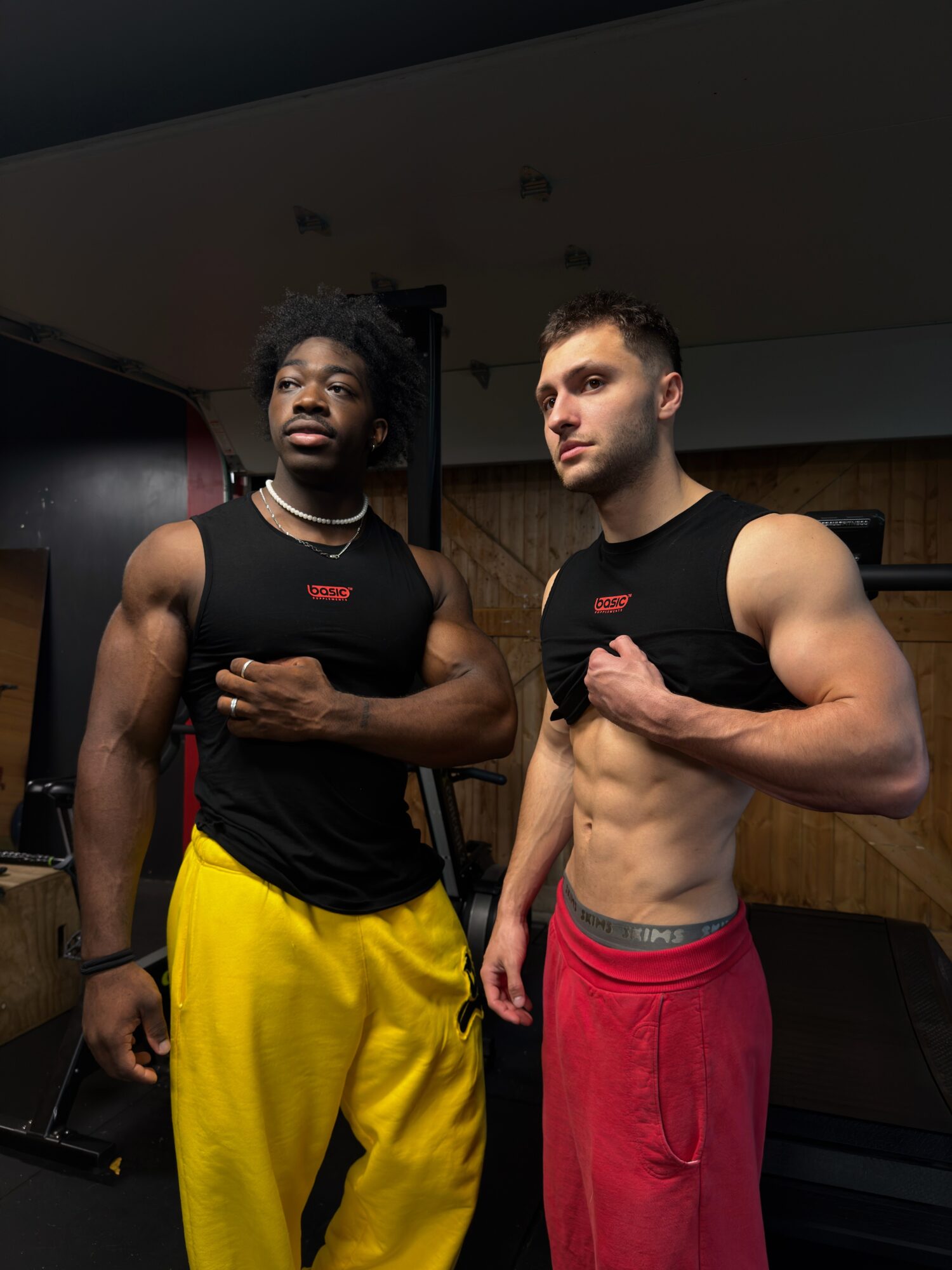 Two muscular men in black tank tops and colorful pants pose indoors, one flexing arm, smiling, with a dark background.