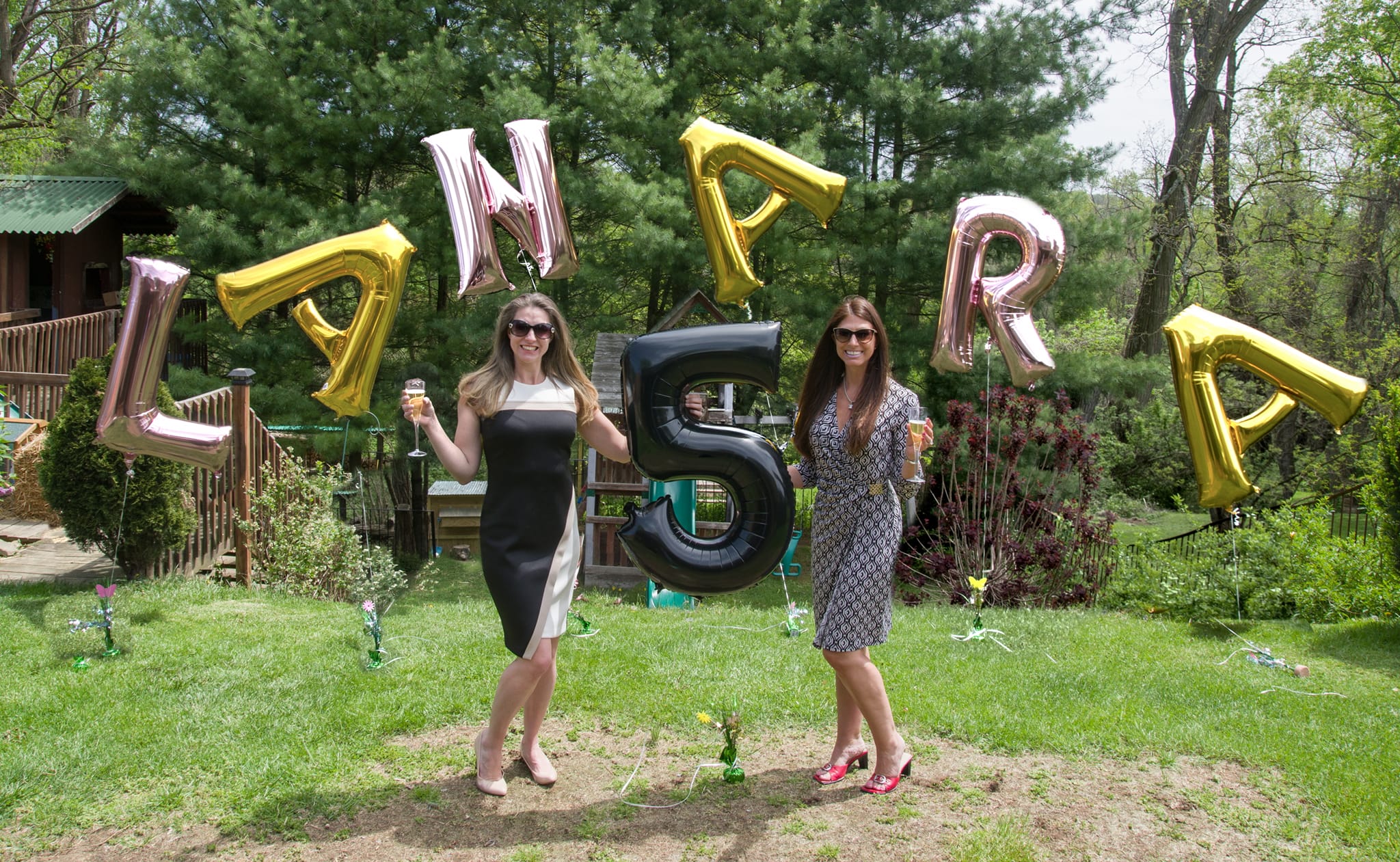 Two women stand outdoors holding balloons spelling 'L', 'A', 'N', 'E', 'R', and a large number '5' balloon, with trees and a house in background.