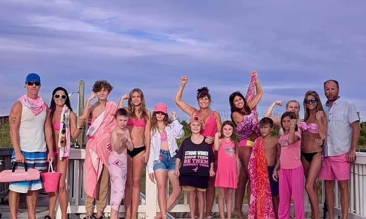Group of people standing outdoors on a beach, some wearing pink clothing, under a cloudy sky.