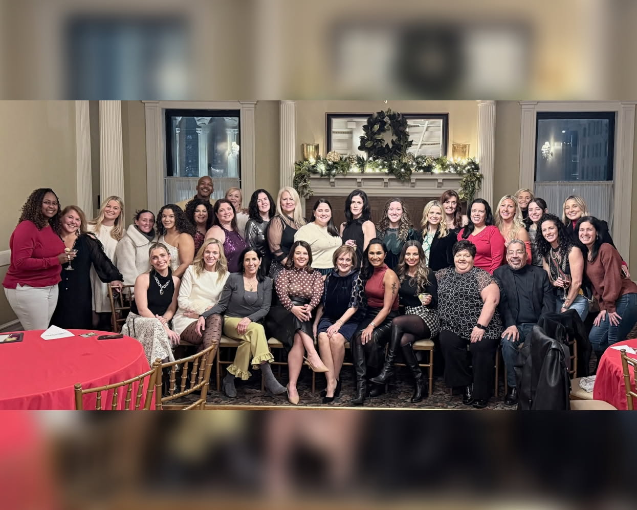 Group of women and men gathered in a decorated room for a celebration, some seated and some standing, smiling at the camera.