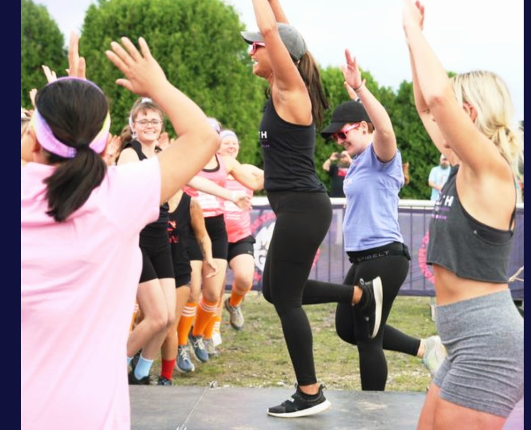 Group of people exercising outdoors, raising arms, with a woman jumping on one leg in the center, trees in background.