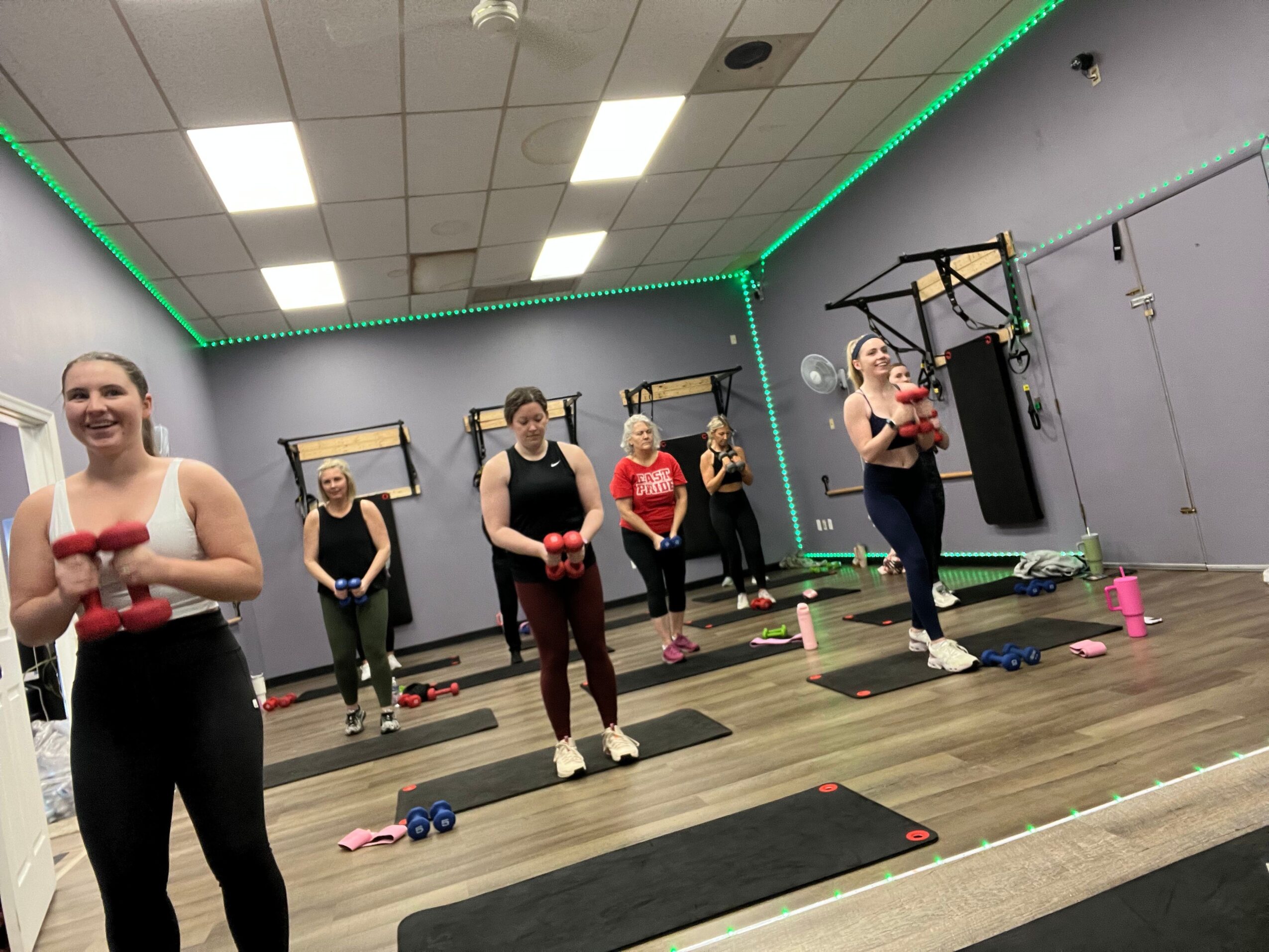 Group of people in a fitness class, standing on mats in a room with gray walls and green LED lighting, facing the camera.