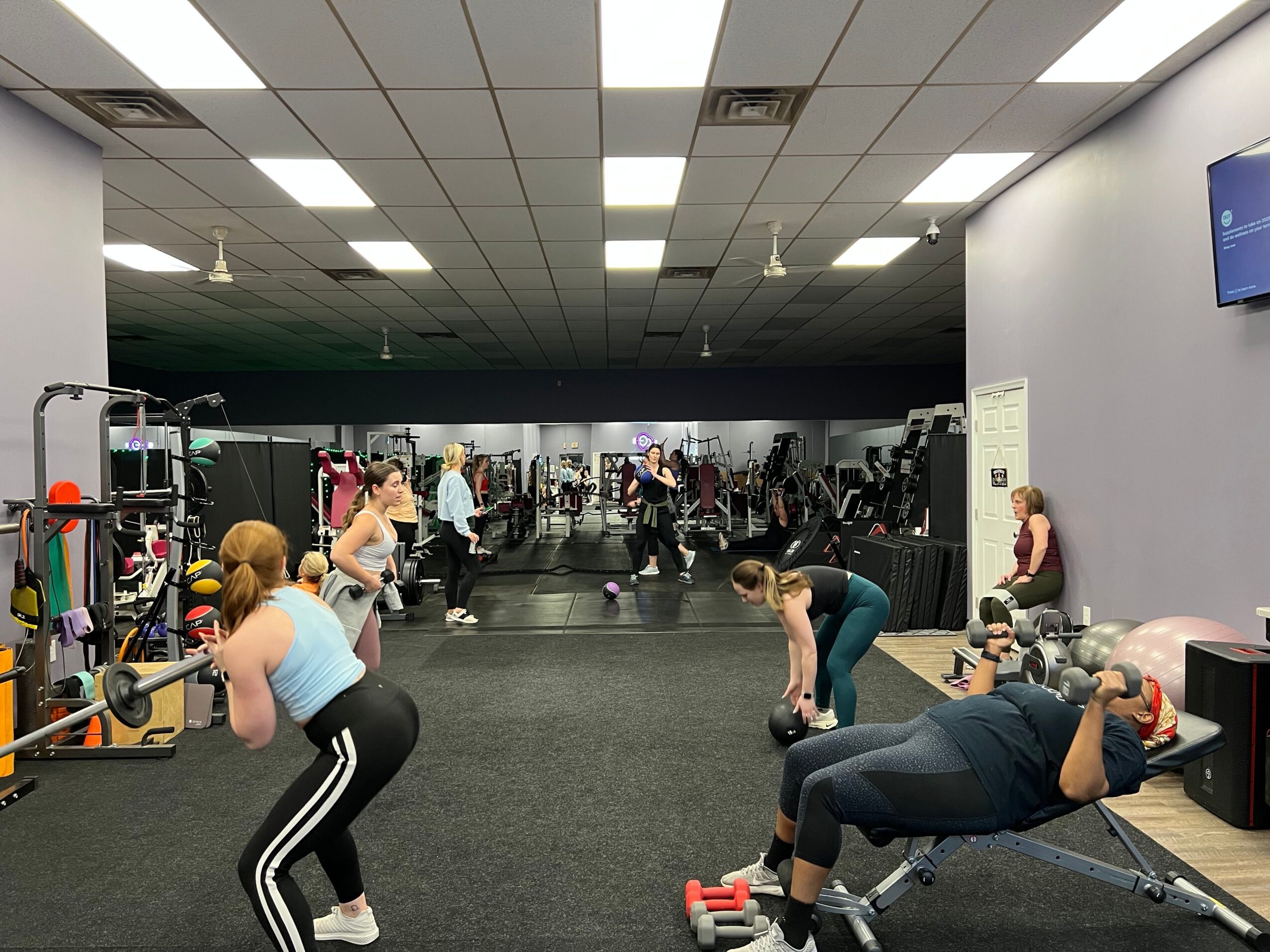 People working out in a gym with exercise equipment and mirrors, some using weights and others stretching.