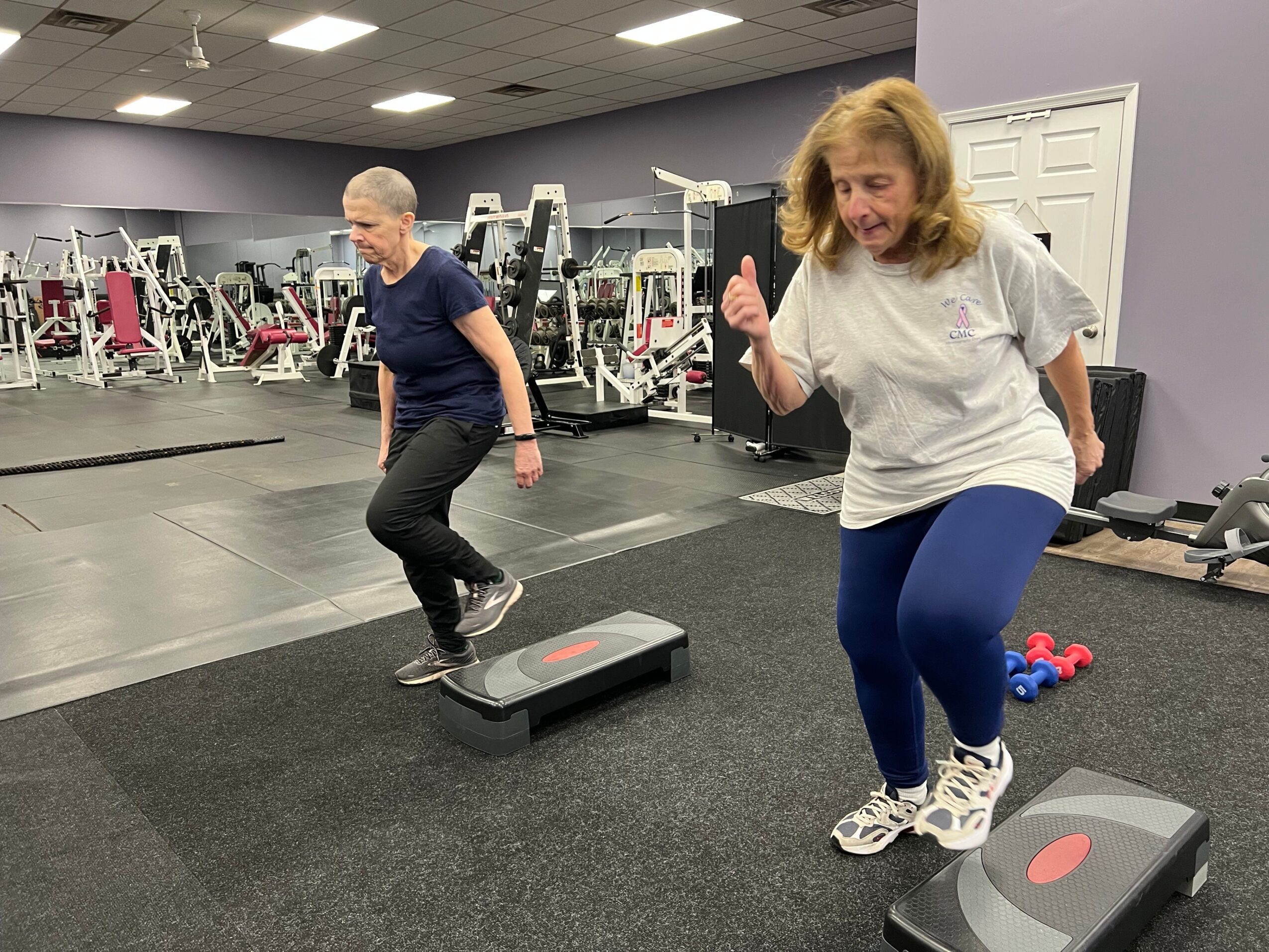 Two women in a gym, one standing and one lying on the floor, with gym equipment in the background.