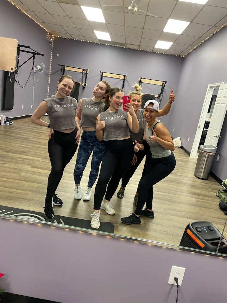 Five women in workout clothes taking a mirror selfie in a gym, smiling and posing cheerfully.