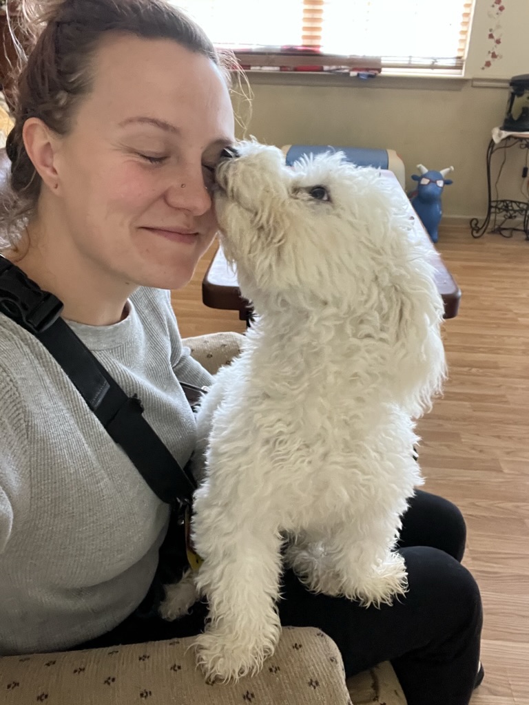 Person with curly hair smiling as a fluffy white puppy licks their face indoors.