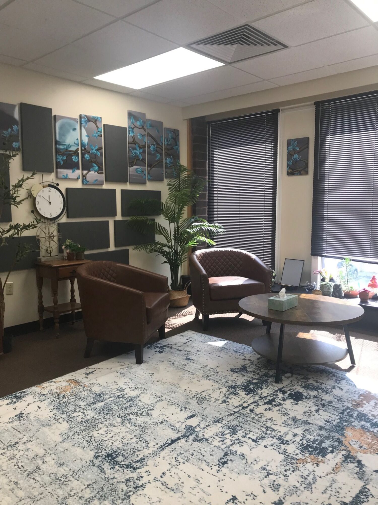 Office with a desk, chair, computer, plant, and wall art, viewed from the side with windows and blinds.