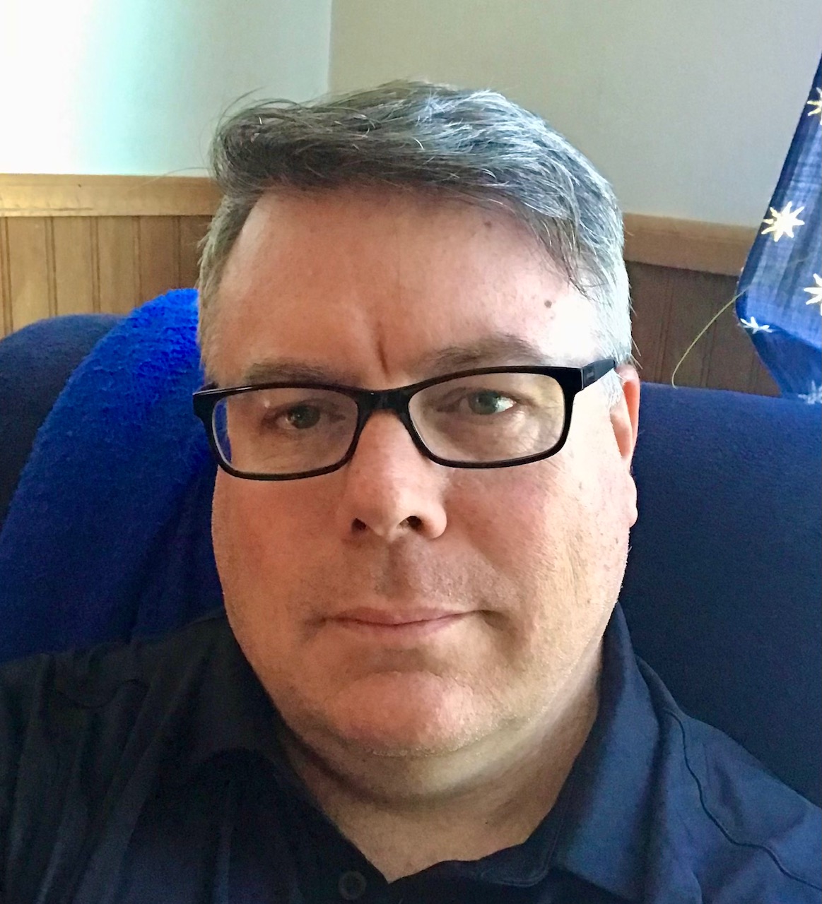 Man with glasses and short gray hair sitting indoors, wearing a dark shirt, with a blue blanket or cushion behind him.