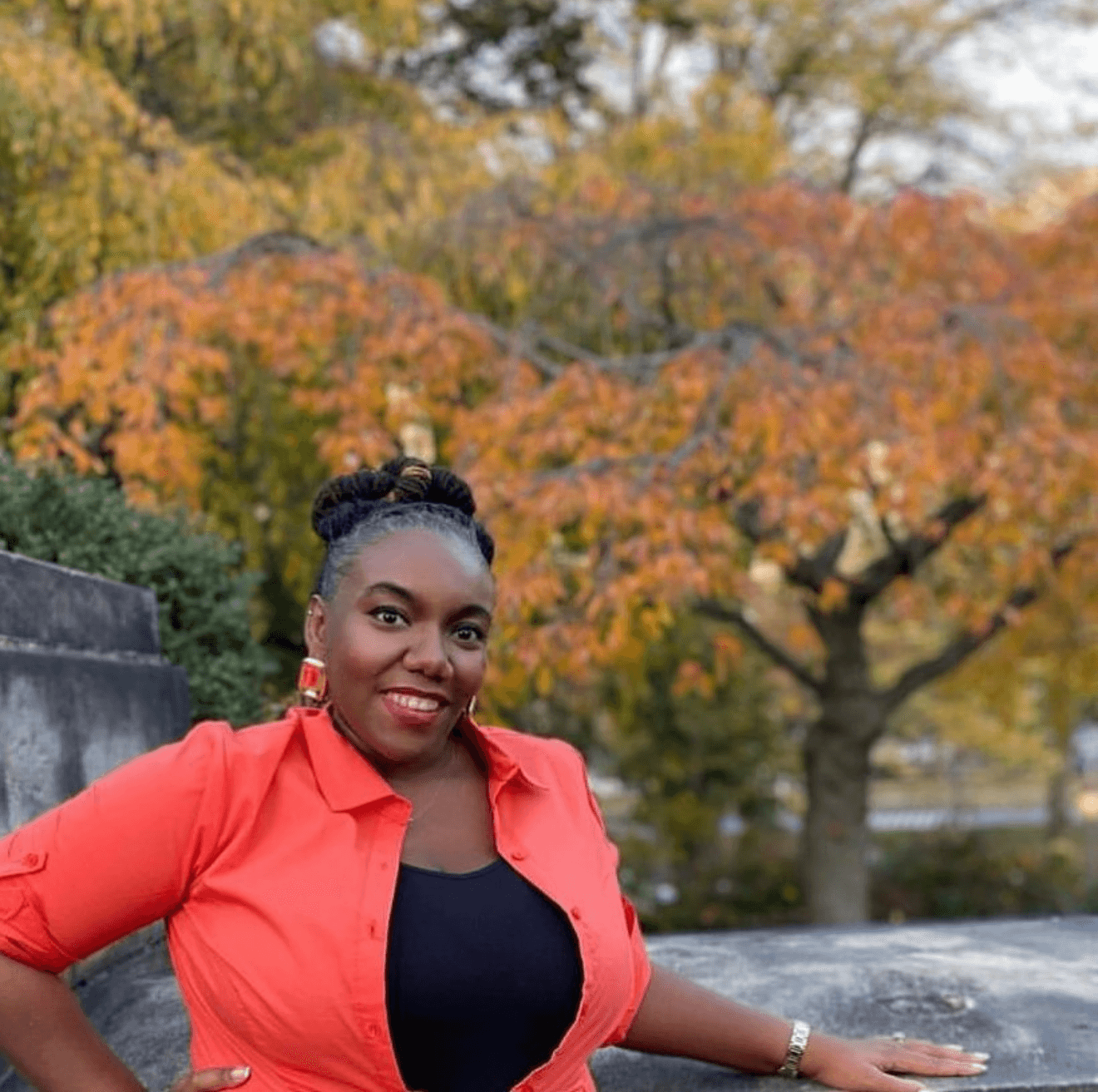 Woman in red shirt outdoors with autumn trees in background, smiling, with arm resting on a stone surface.