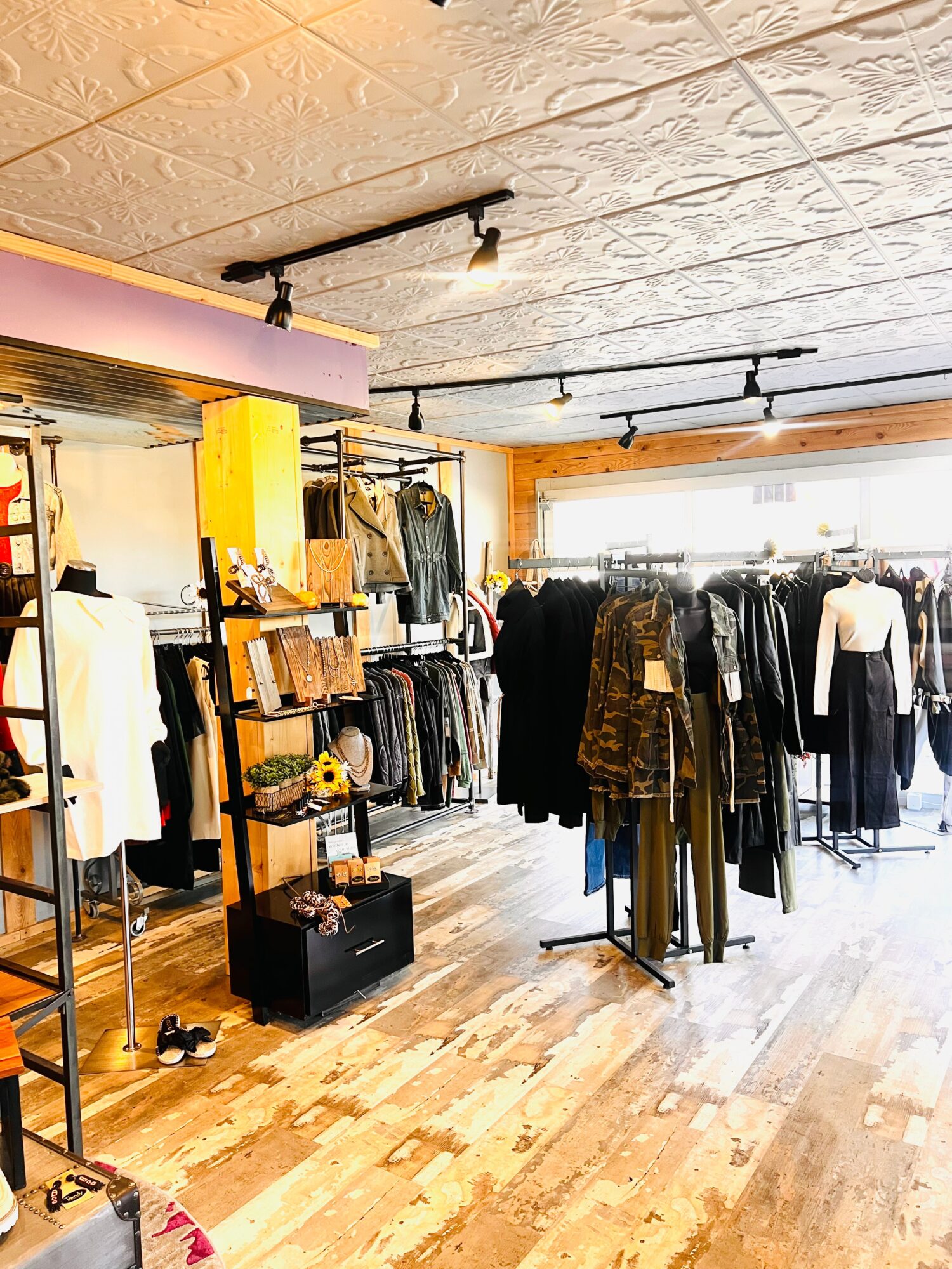 Clothing store interior with racks of clothes, shelves, and a window with sunlight, wooden floor, ceiling lights, and display items.