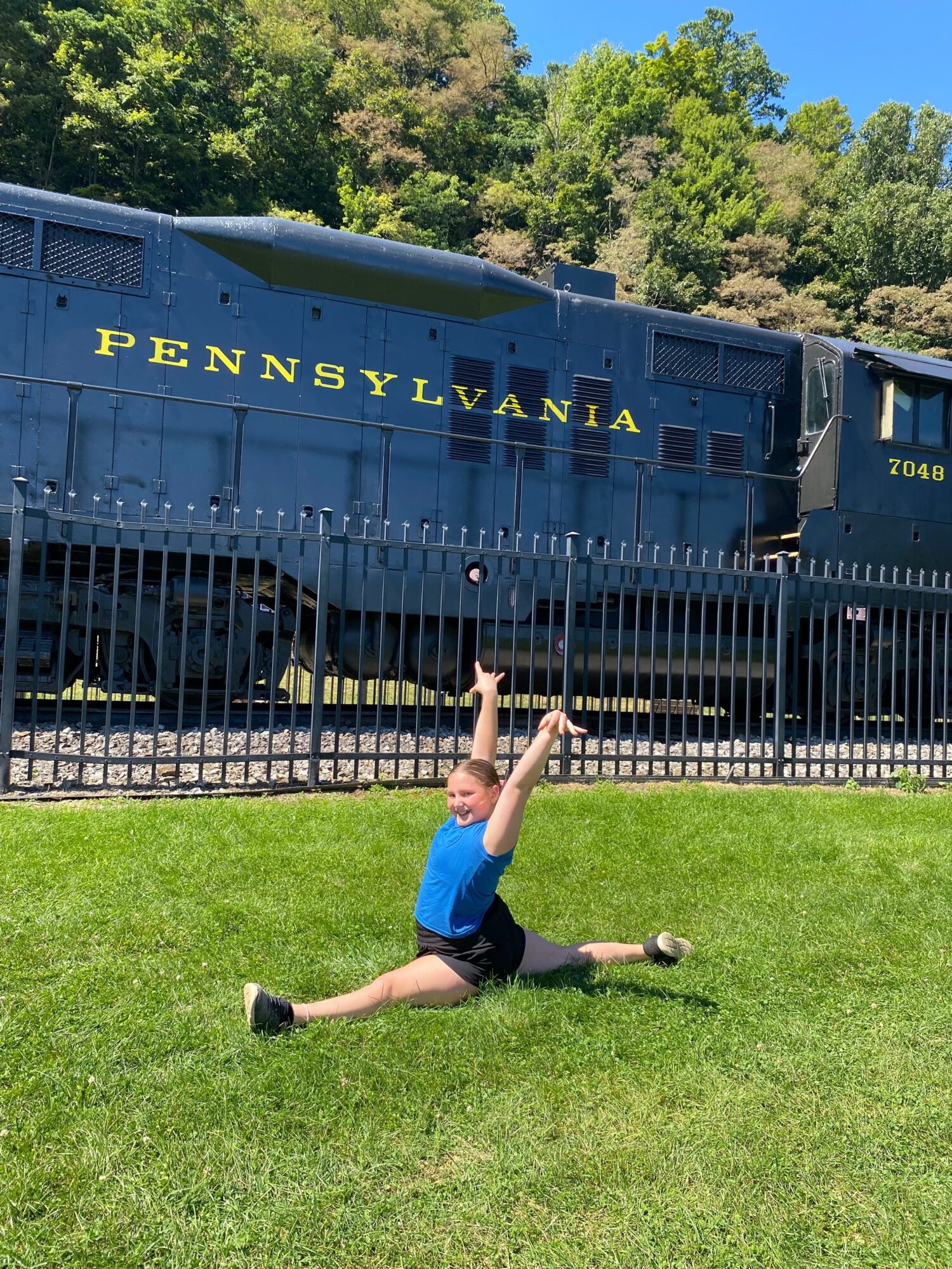 Person lying on grass with arms and legs spread, near a historic train, green trees in background.