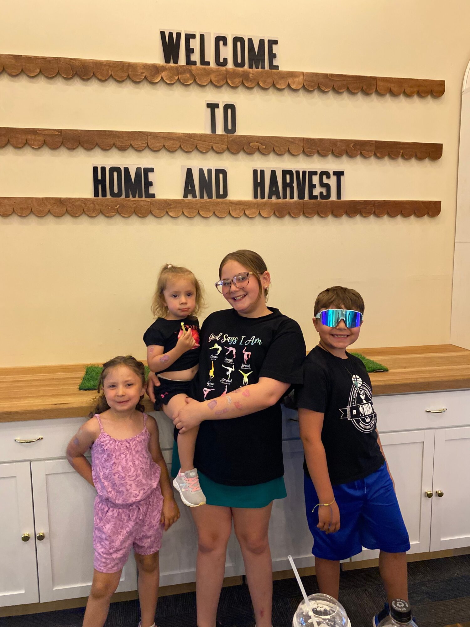 Four children stand in front of a wall with a sign that reads 'Welcome to Home and Harvest.'