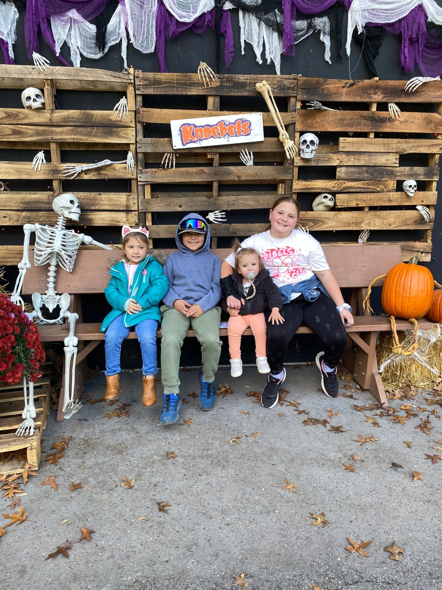 Four children sitting on a wooden pallet display, with pumpkins and decorations around, outdoors on a concrete surface.