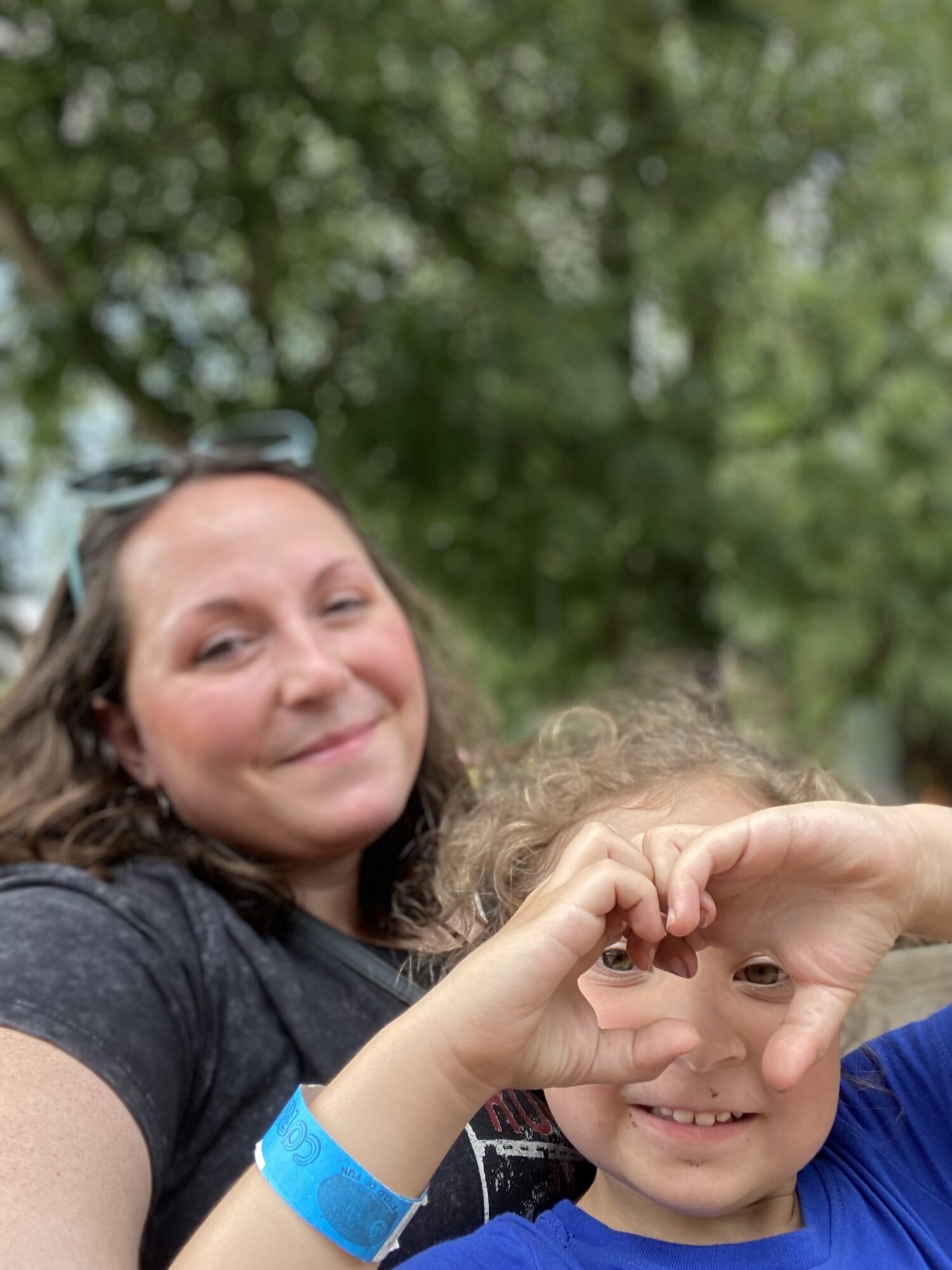 Two smiling people outdoors, one with curly hair and a blue wristband, the other with long hair, making a heart shape with their hand.