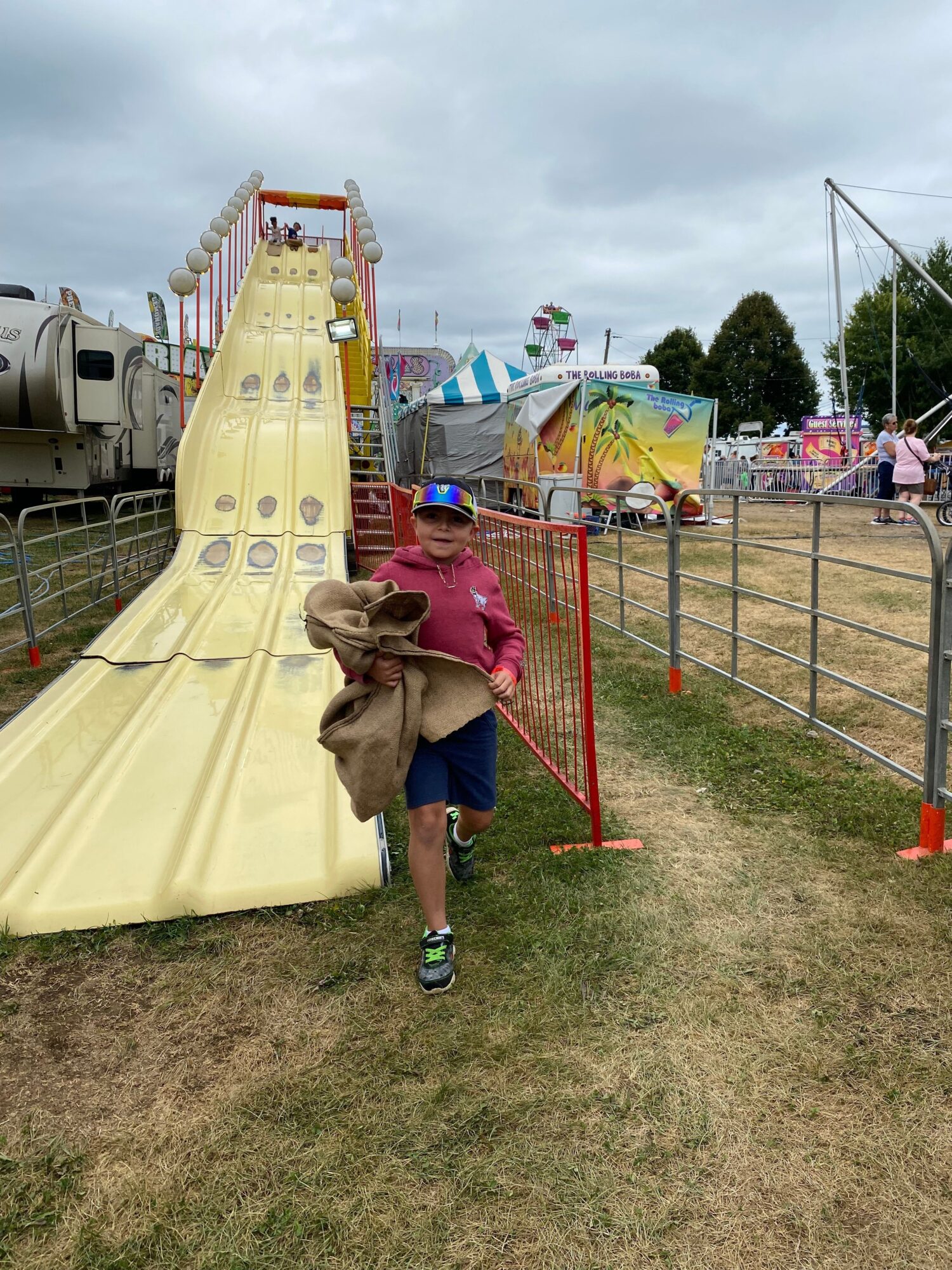Child in pink jacket and blue pants slides down a yellow slide at an outdoor fairground.