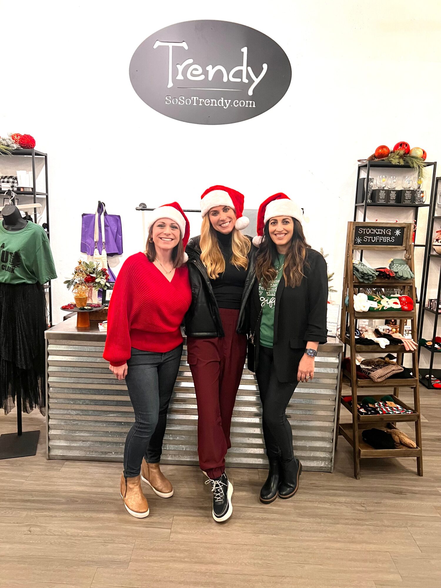 Three women wearing Santa hats and festive clothing stand inside a store with shelves of clothing and accessories behind them.
