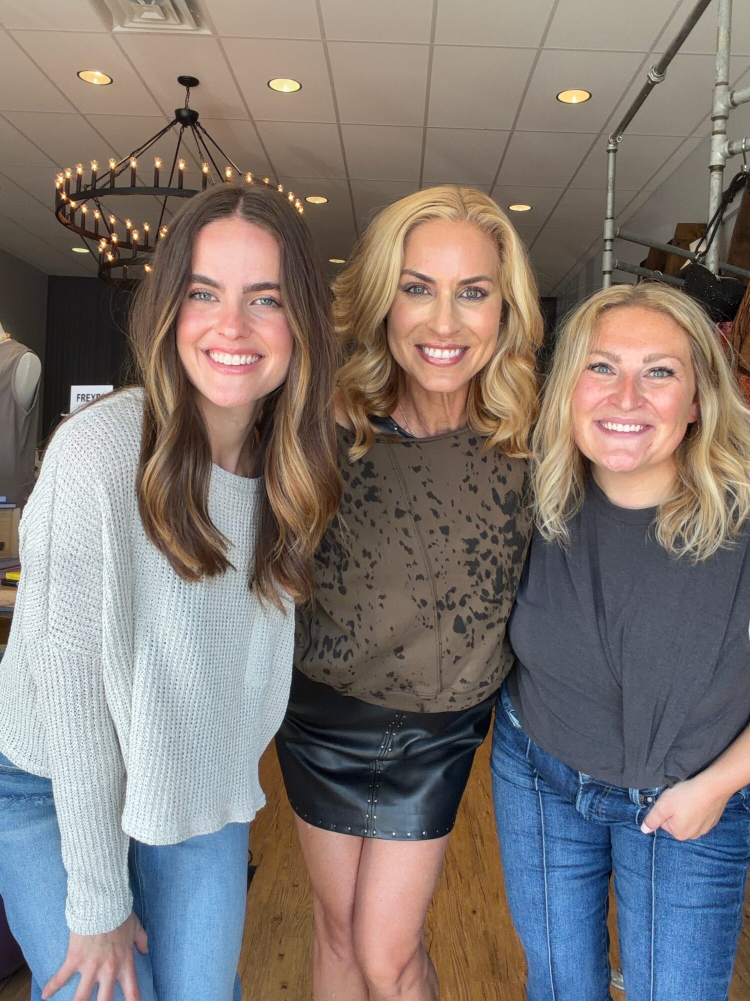 Three women smiling indoors, chandelier hanging from ceiling, wooden floor, casual clothing, close together.