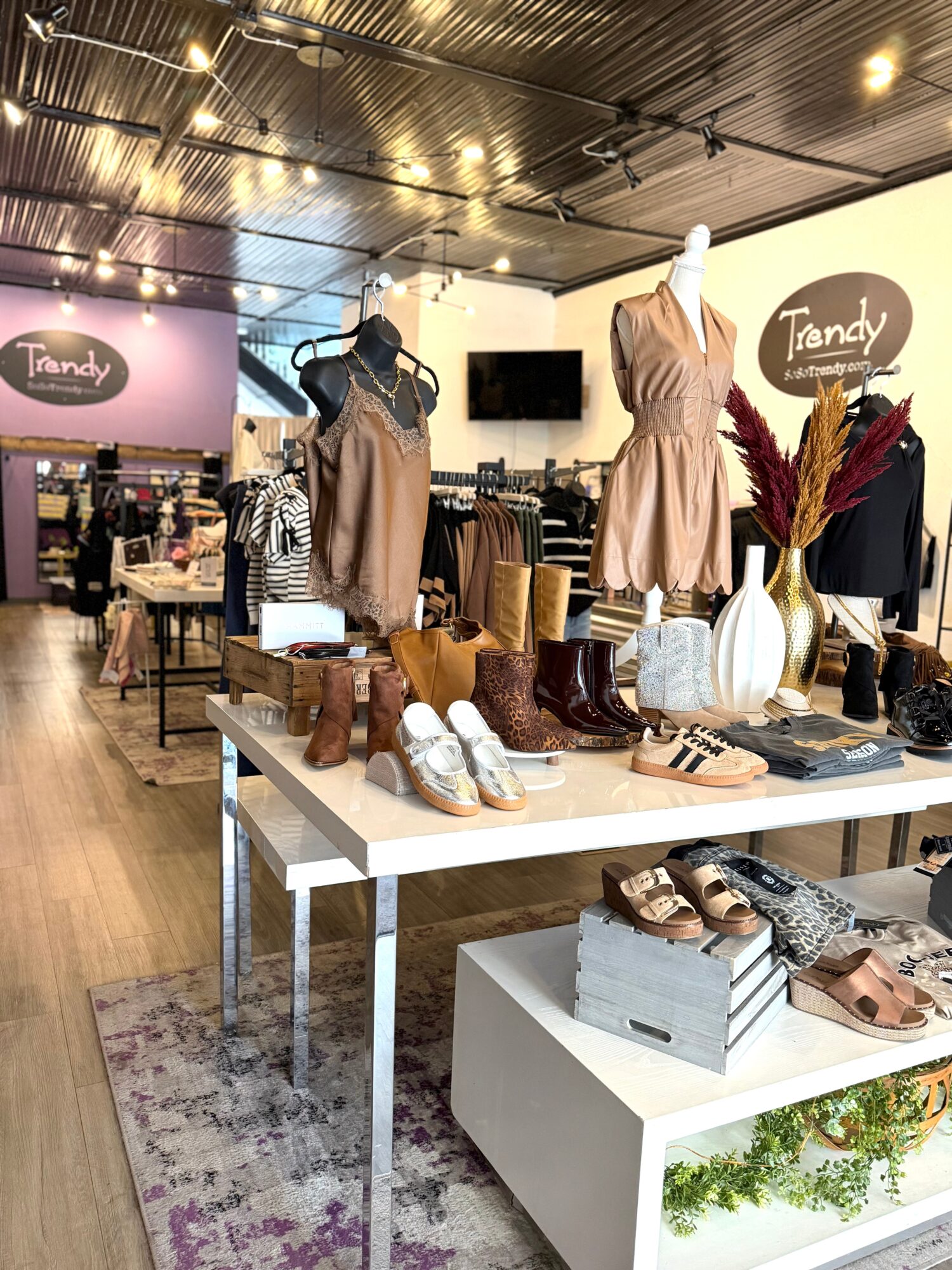 Clothing store display with mannequins wearing dresses, shoes, and accessories on tables, store interior visible in background.