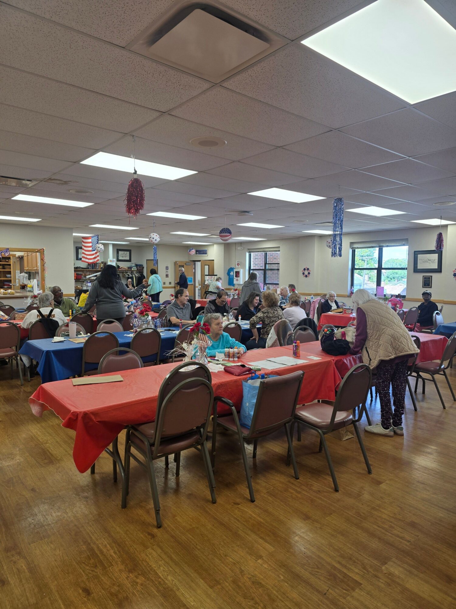 People gathered around tables with red, blue, and white tablecloths in a room with wooden floors and large windows.