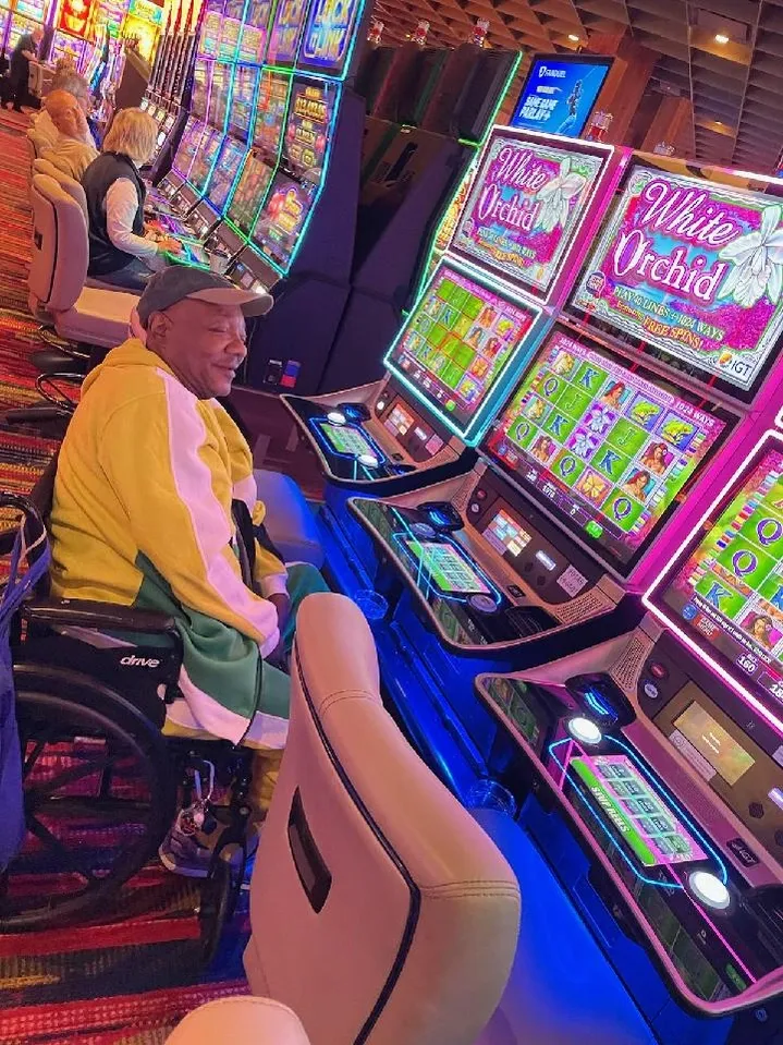 Person in a yellow jacket sitting at a slot machine in a casino with colorful gaming screens, other players in background.