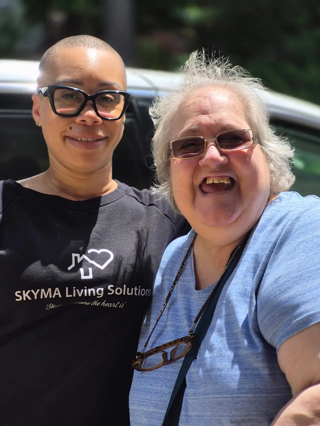 Two women smiling outdoors, one with glasses and short hair, the other with glasses and gray hair, in front of a car.
