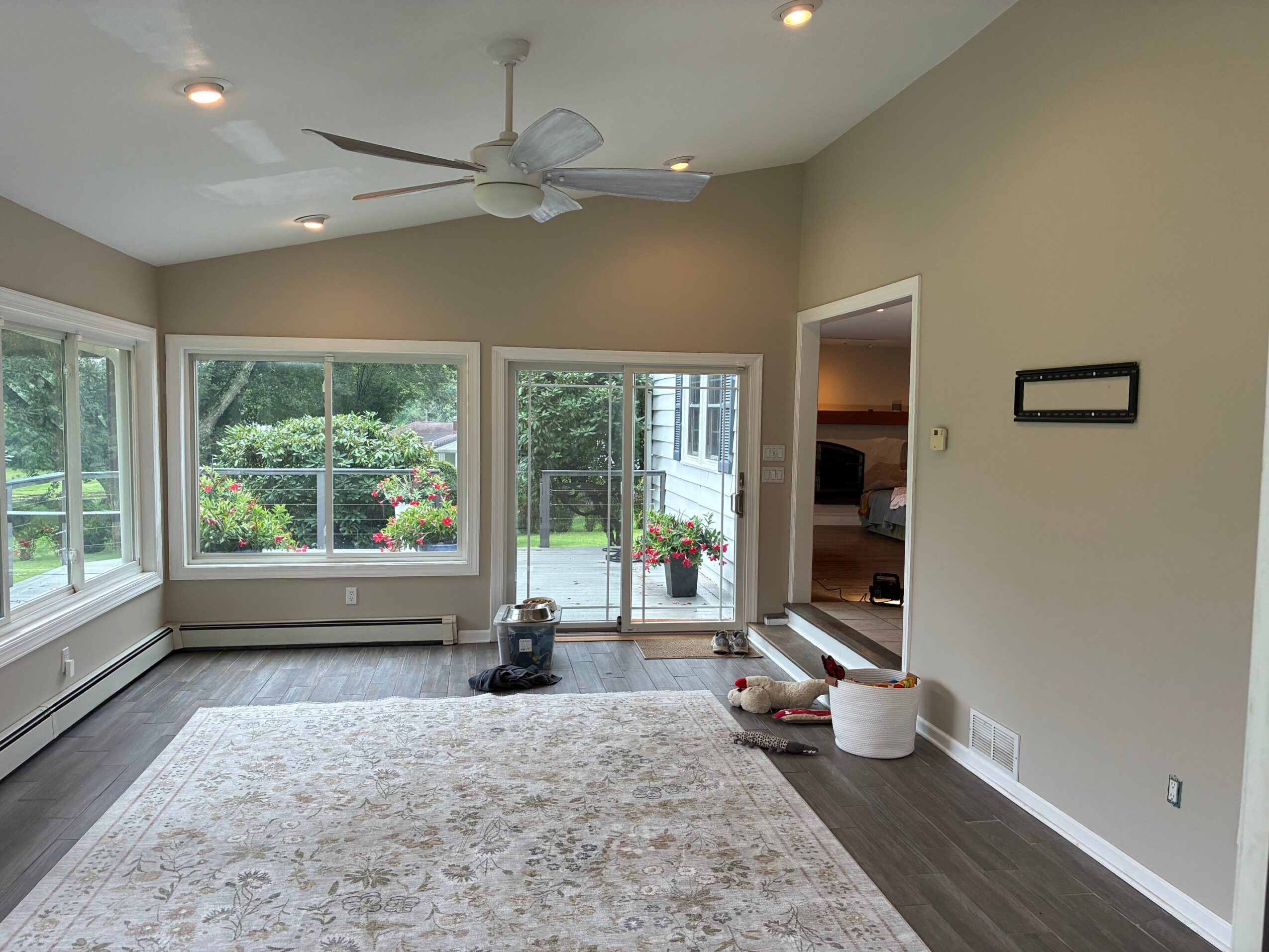 Living room with large windows, ceiling fan, and hardwood floor, with a door leading outside and some objects near the wall.