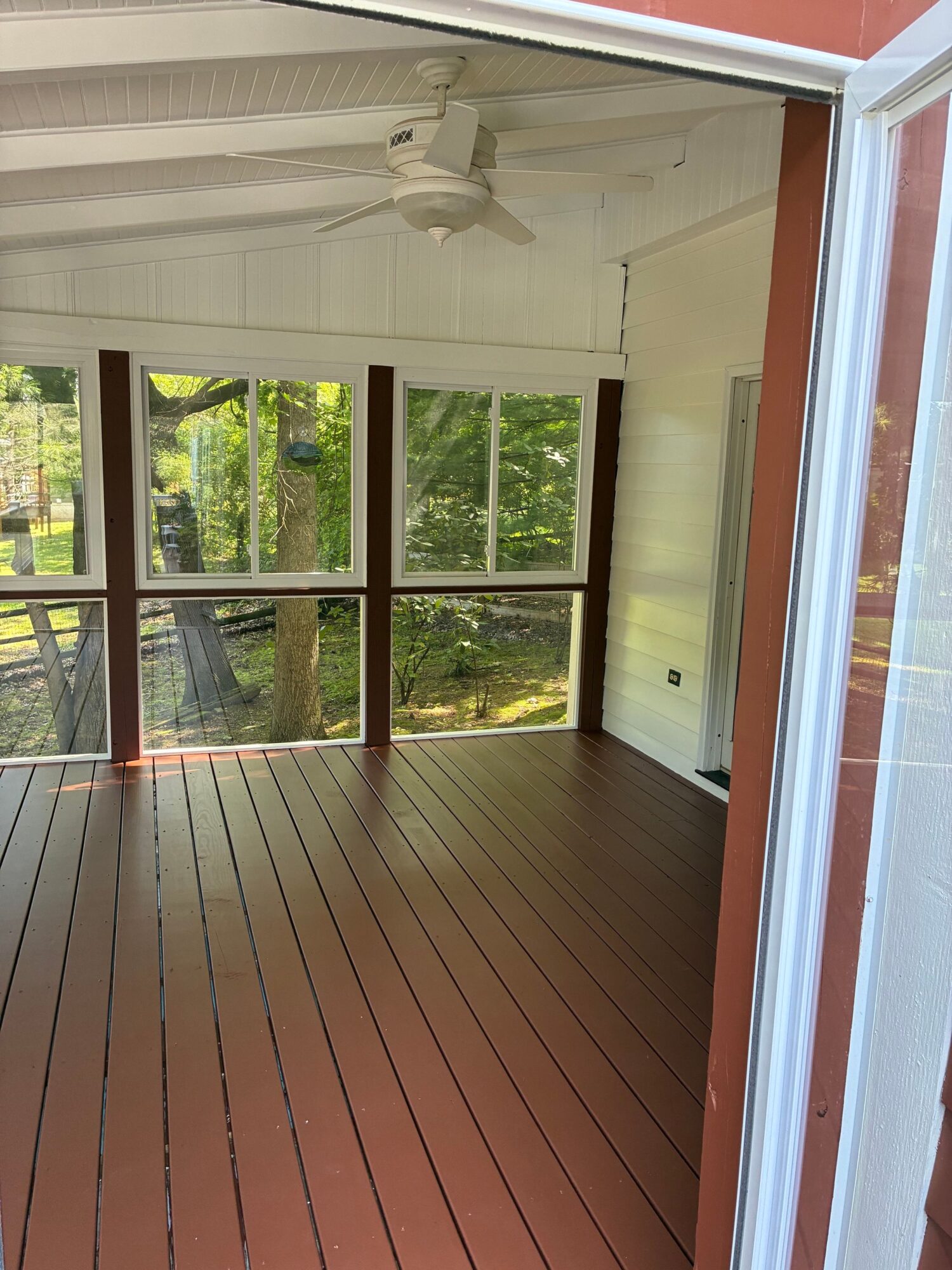View from a porch into a room with large windows showing green trees outside, wooden floor, ceiling fan, and white walls.