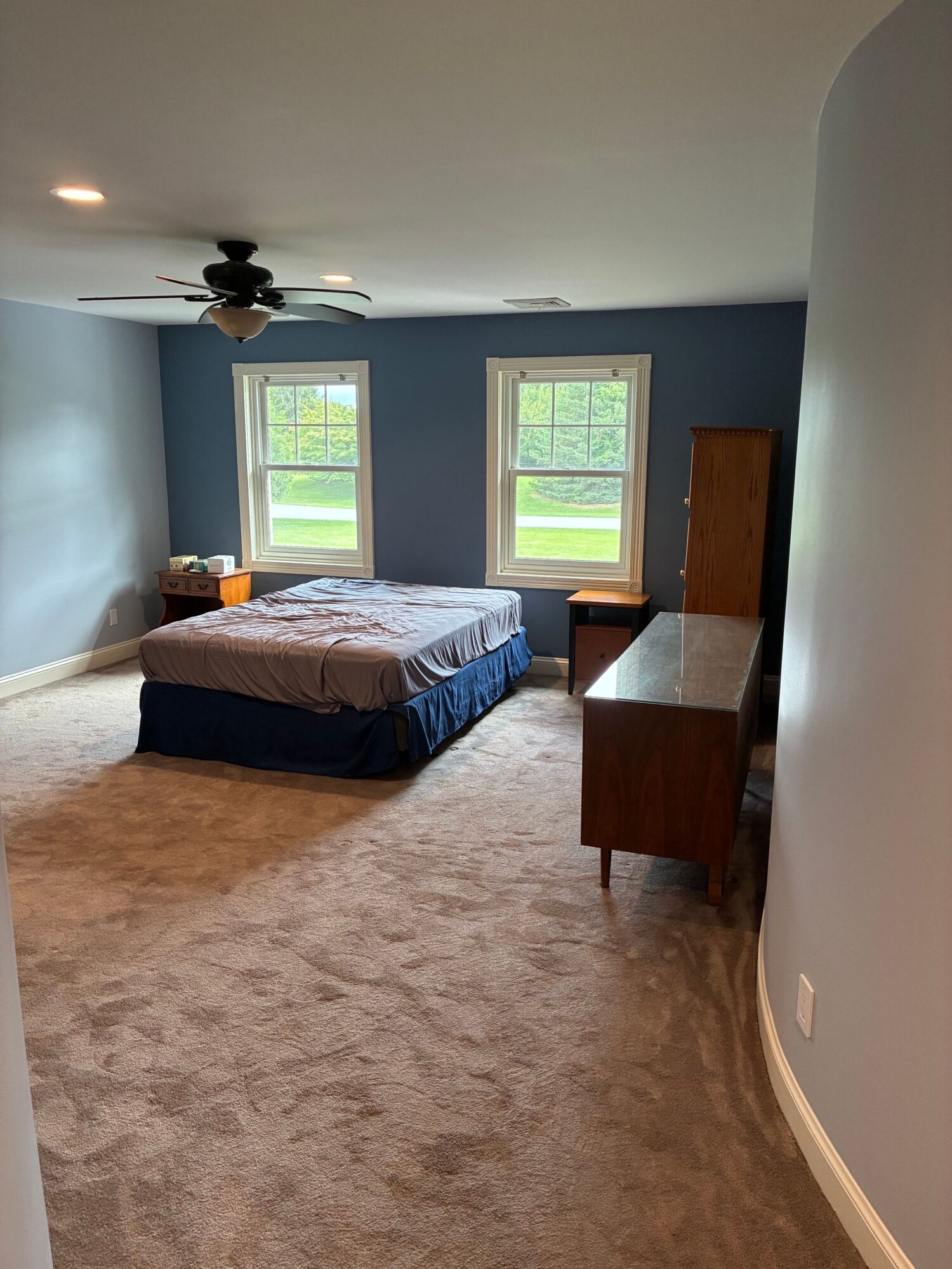 Bedroom with two windows, bed, wooden dresser, and ceiling fan, carpeted floor, painted walls, and natural light.