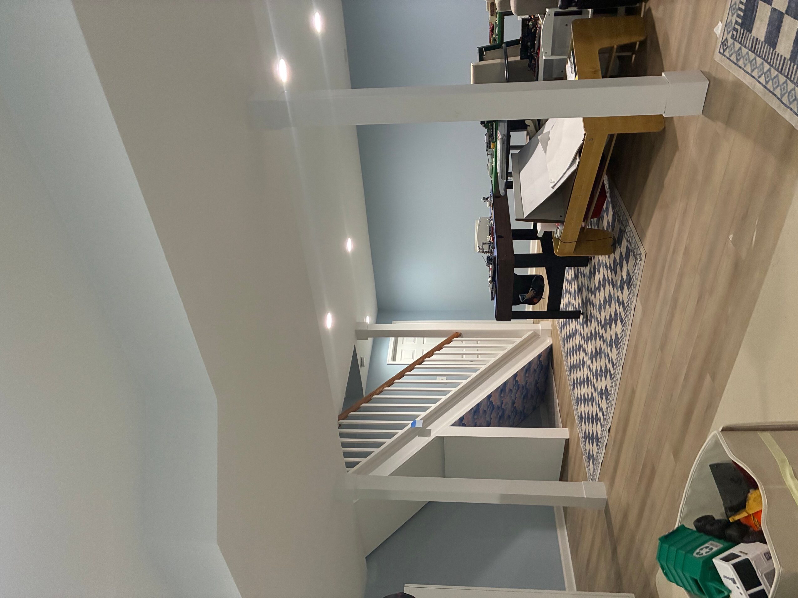 Living room with a dining table, chairs, and a staircase, viewed from the side with ceiling lights and wooden flooring.