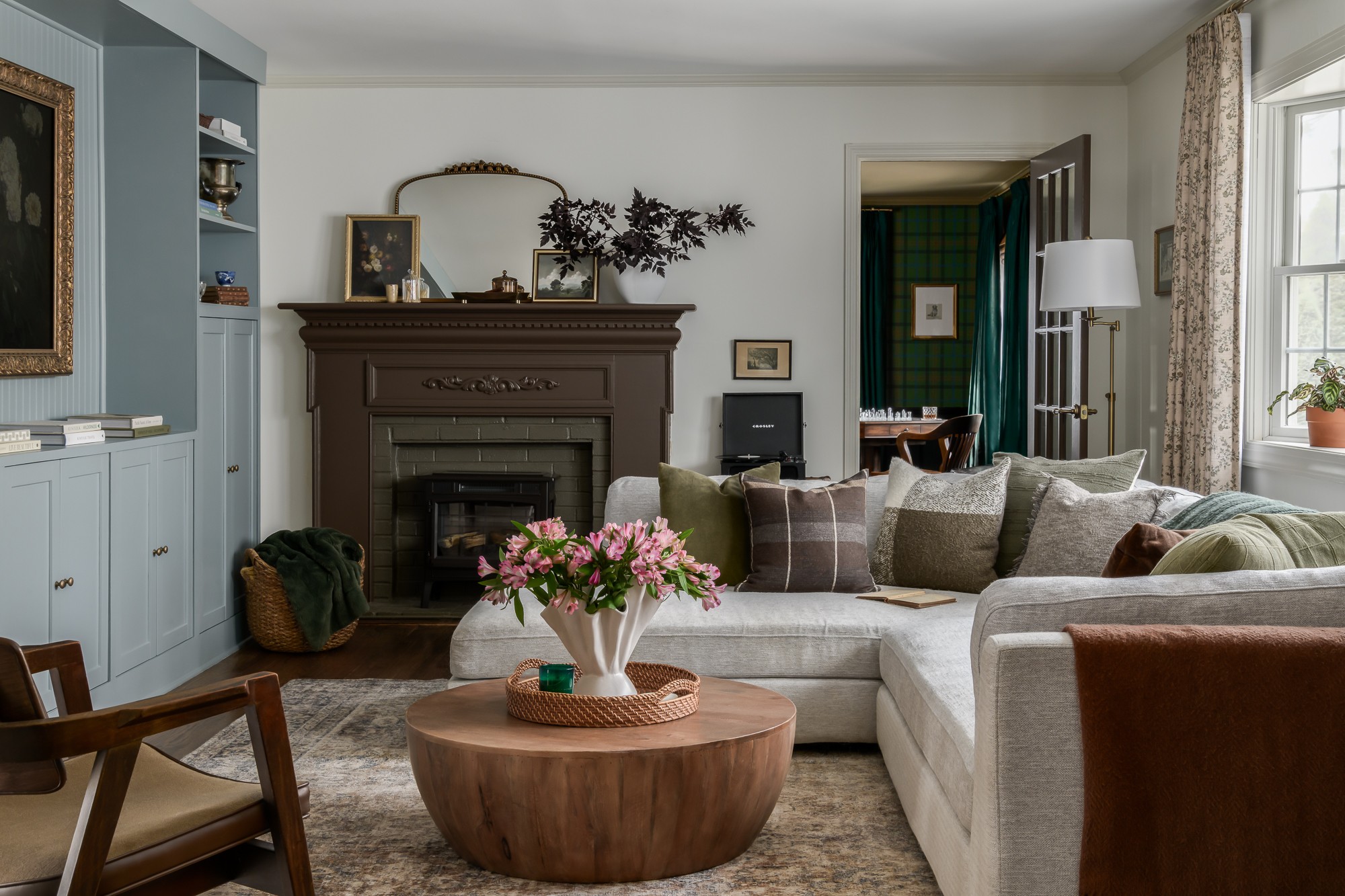 Living room with a fireplace, sofa, armchair, and a round wooden coffee table with a flower vase, bright window, and decorative items.