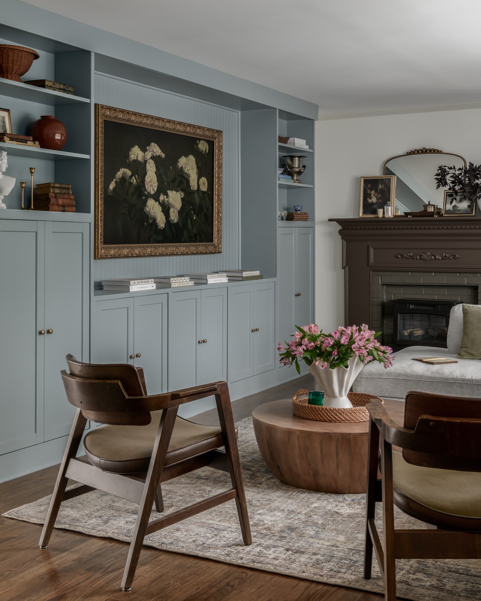 Living room with blue built-in shelves, a fireplace, a large painting, two chairs, a round wooden table with pink flowers, and a rug.