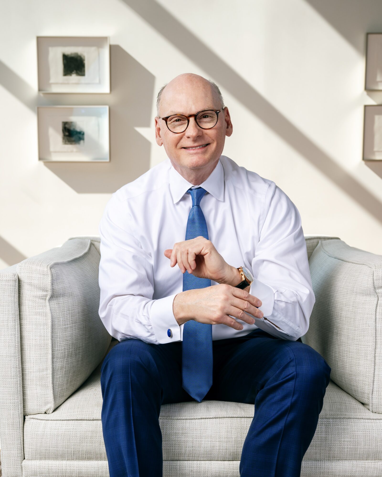 Smiling man with glasses in a white shirt and blue tie sitting on a light-colored sofa in a bright room.