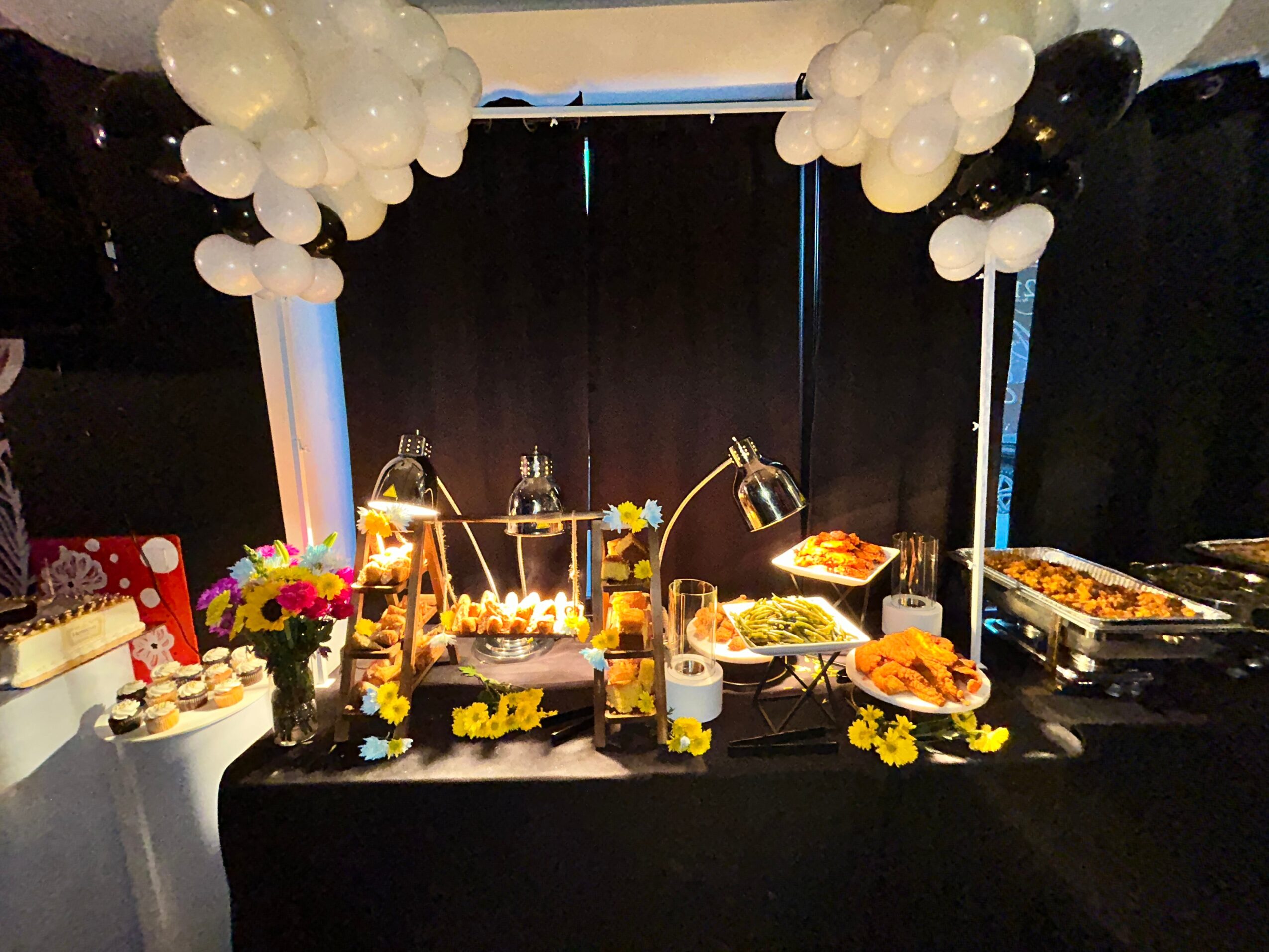 Dessert table with balloons, flowers, and various sweet treats, set against a dark curtain backdrop.