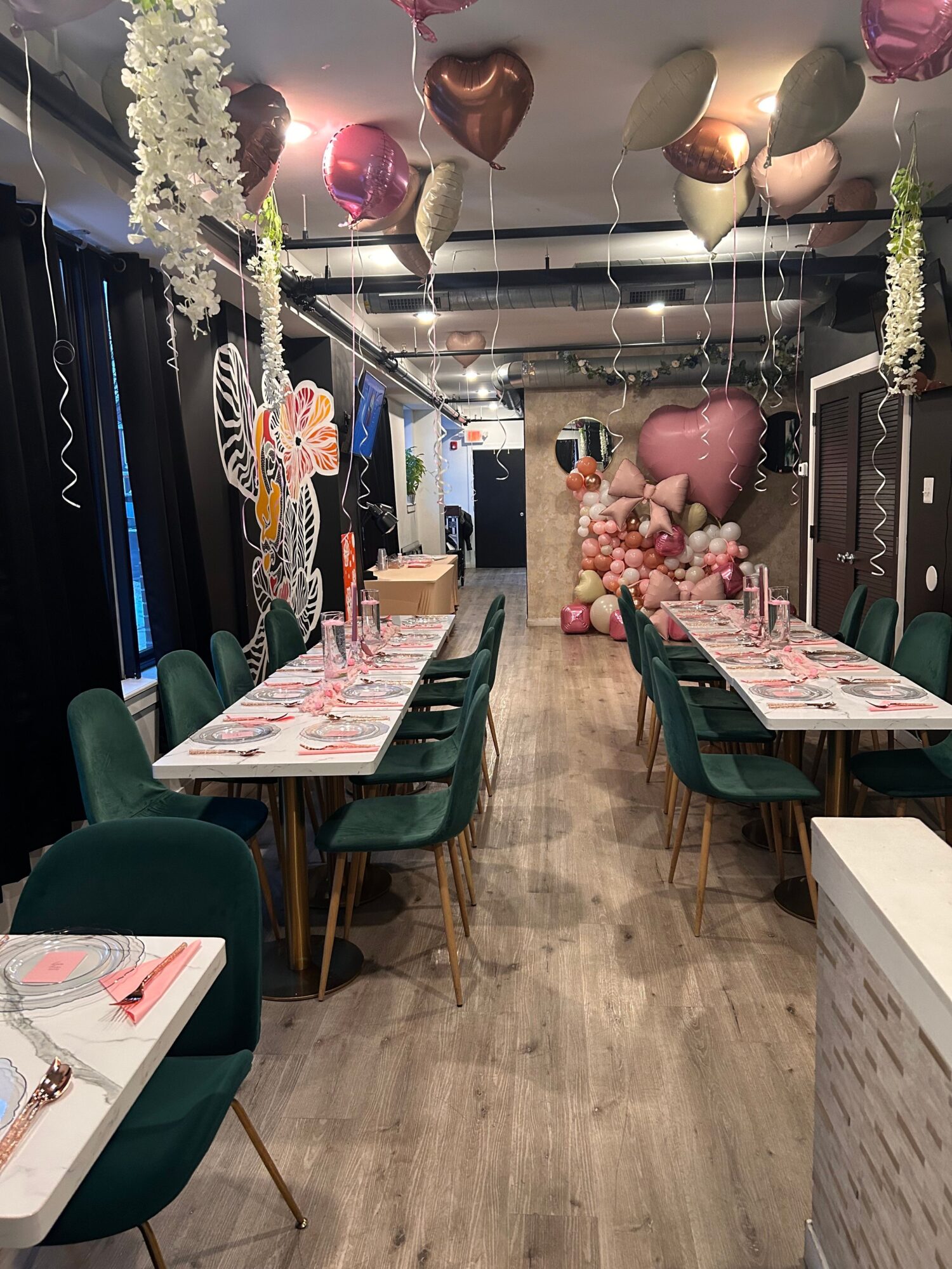 Party setup with balloons, decorations, and tables with pink and white tablecloths, chairs, and floral arrangements.