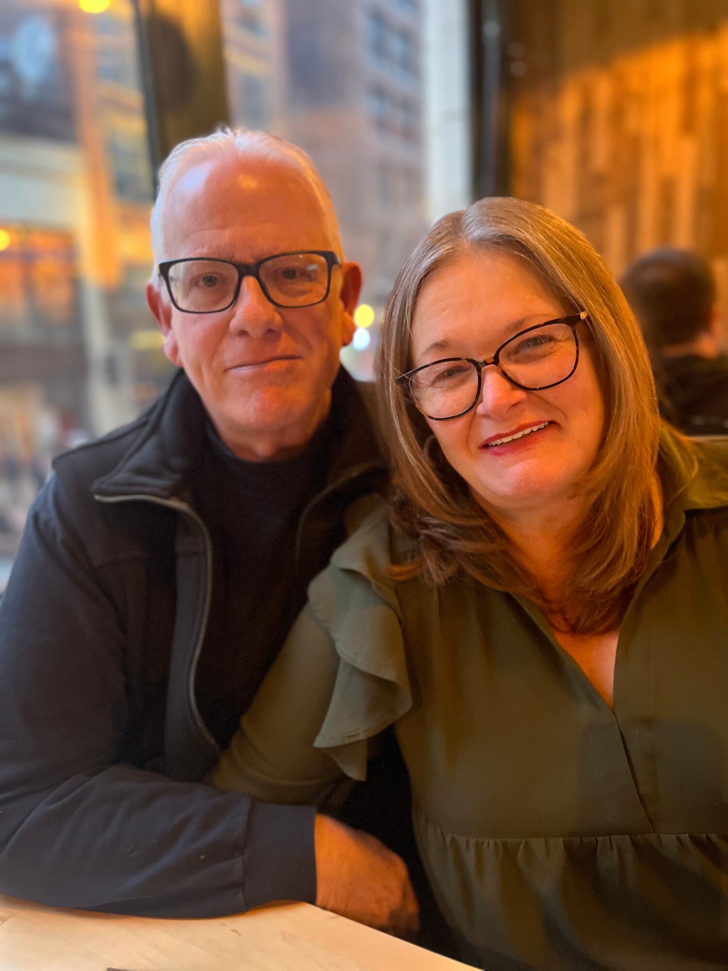 Two people sitting together indoors, smiling, with city lights visible through windows behind them.