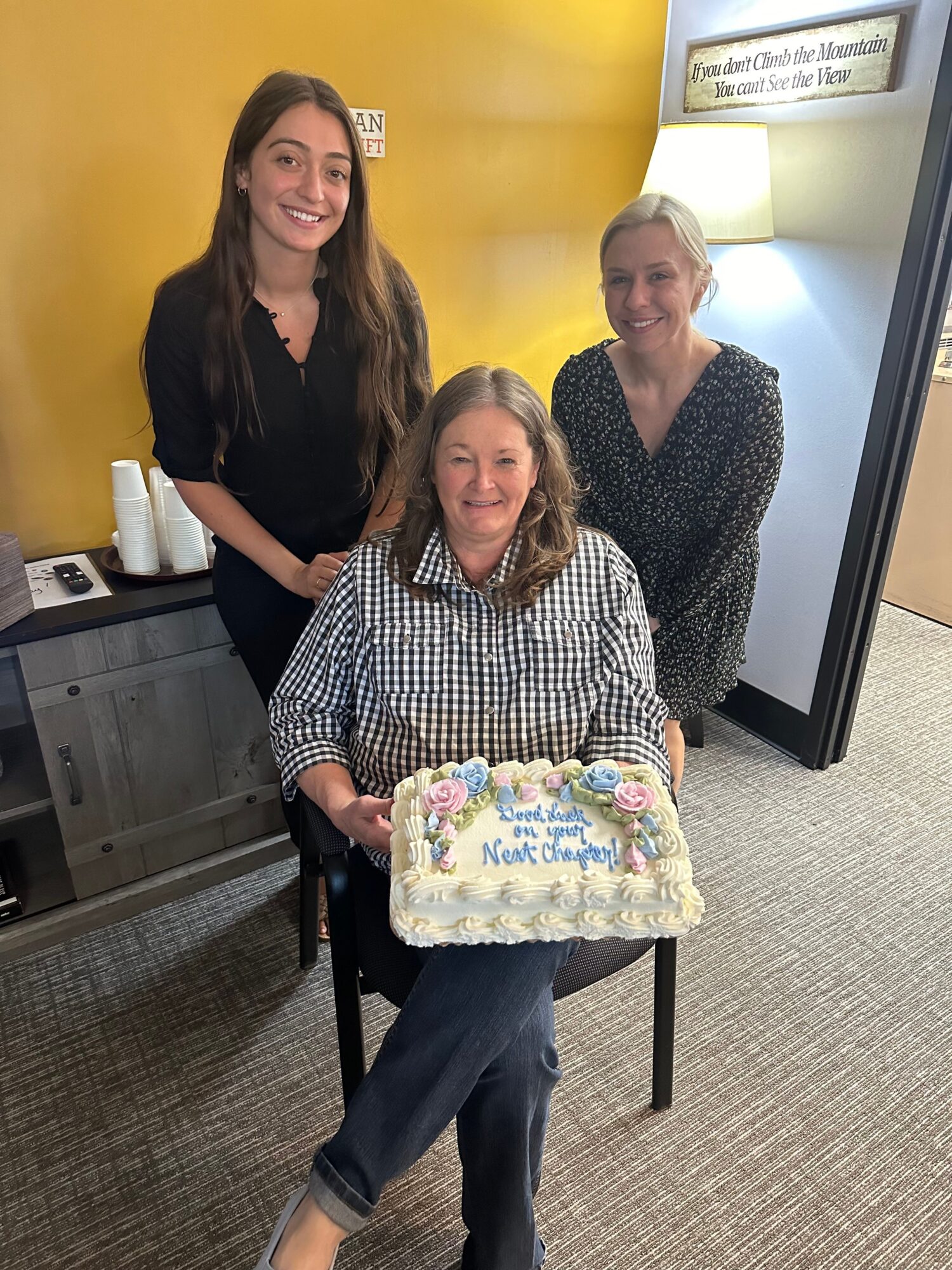 Three women in an office, one seated with a cake, two standing behind, smiling, with a yellow wall and lamp in background.
