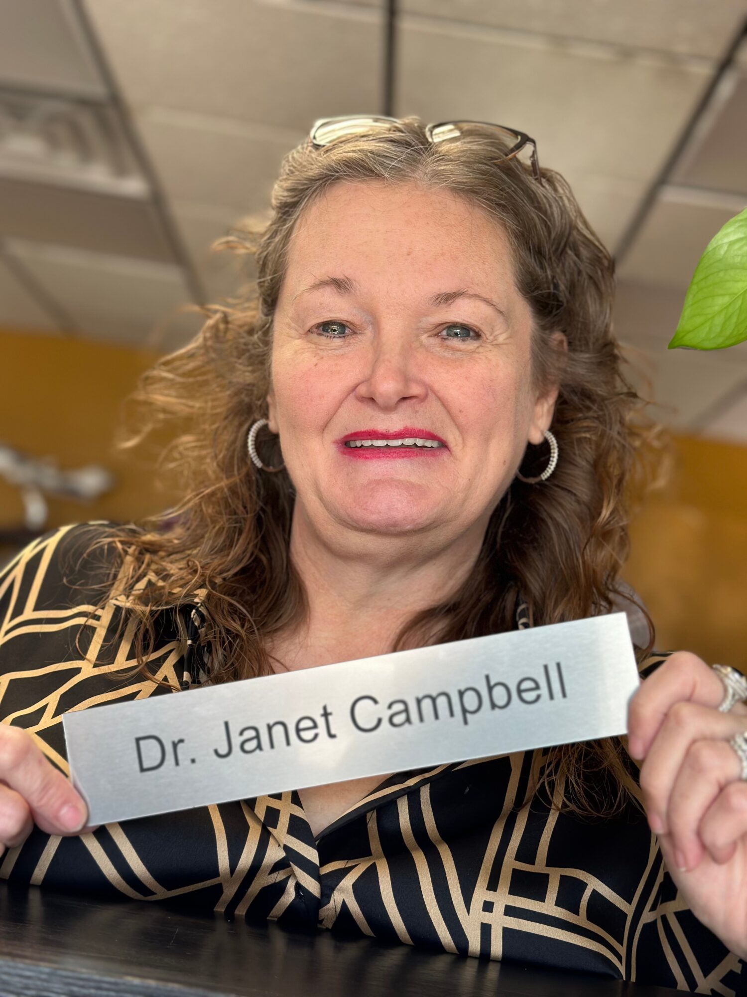 Woman holding a sign with her name, smiling, in an indoor setting with ceiling tiles and a plant visible.