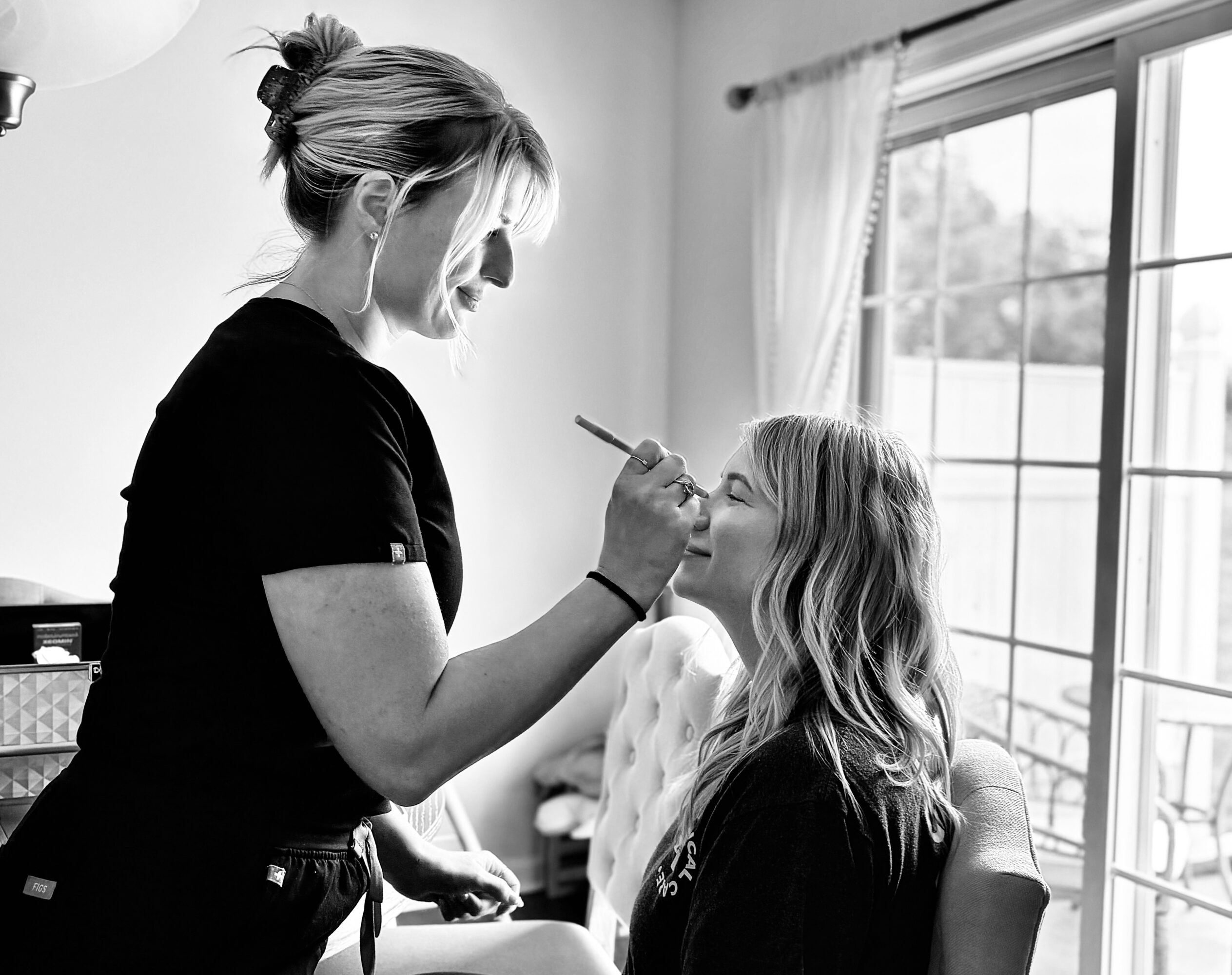 Woman applying makeup to a seated girl near a window with curtains, in a bright room.