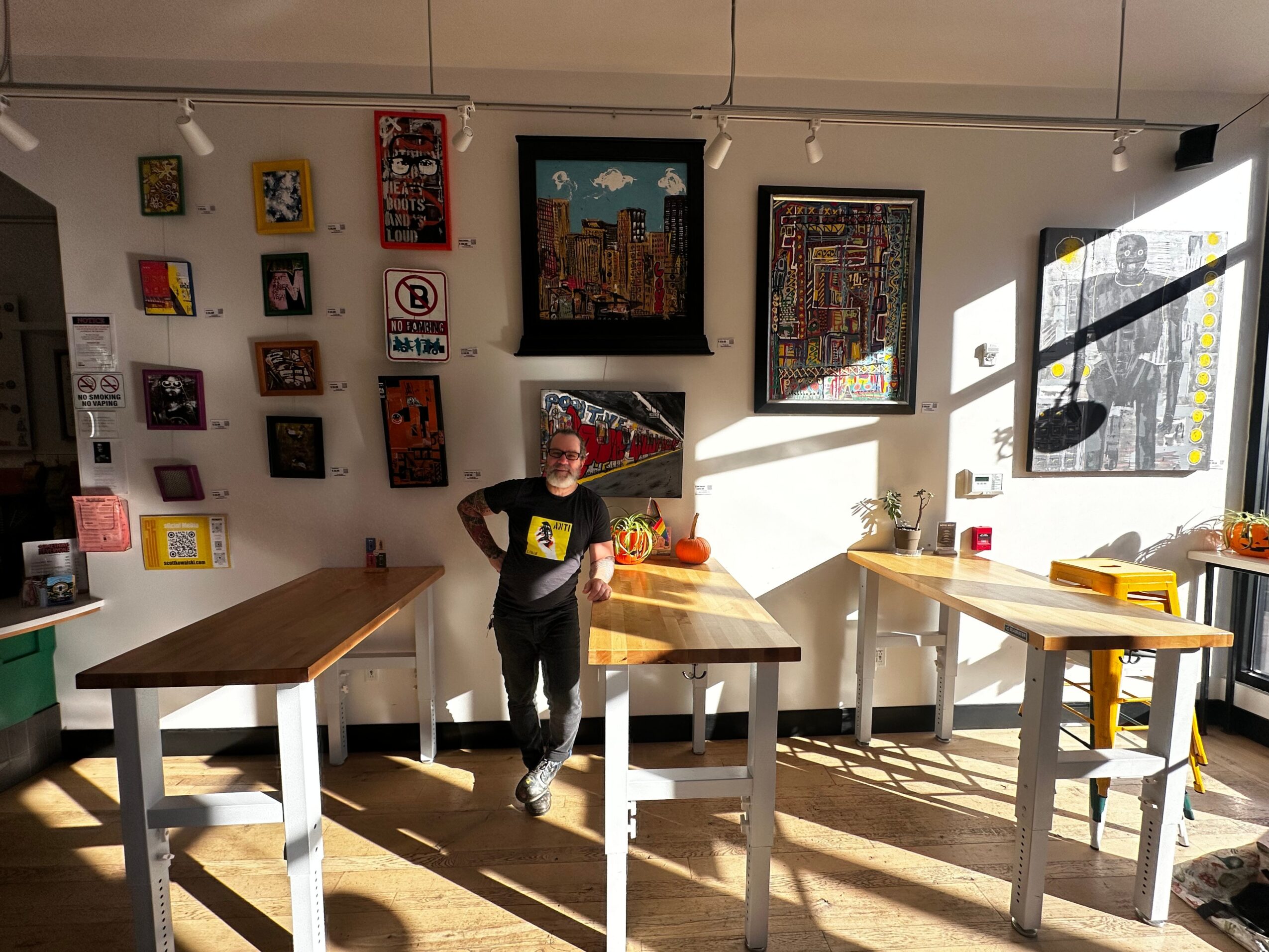 Child standing in art gallery with colorful framed artworks on white wall, sunlight streaming through window, shadows on floor.