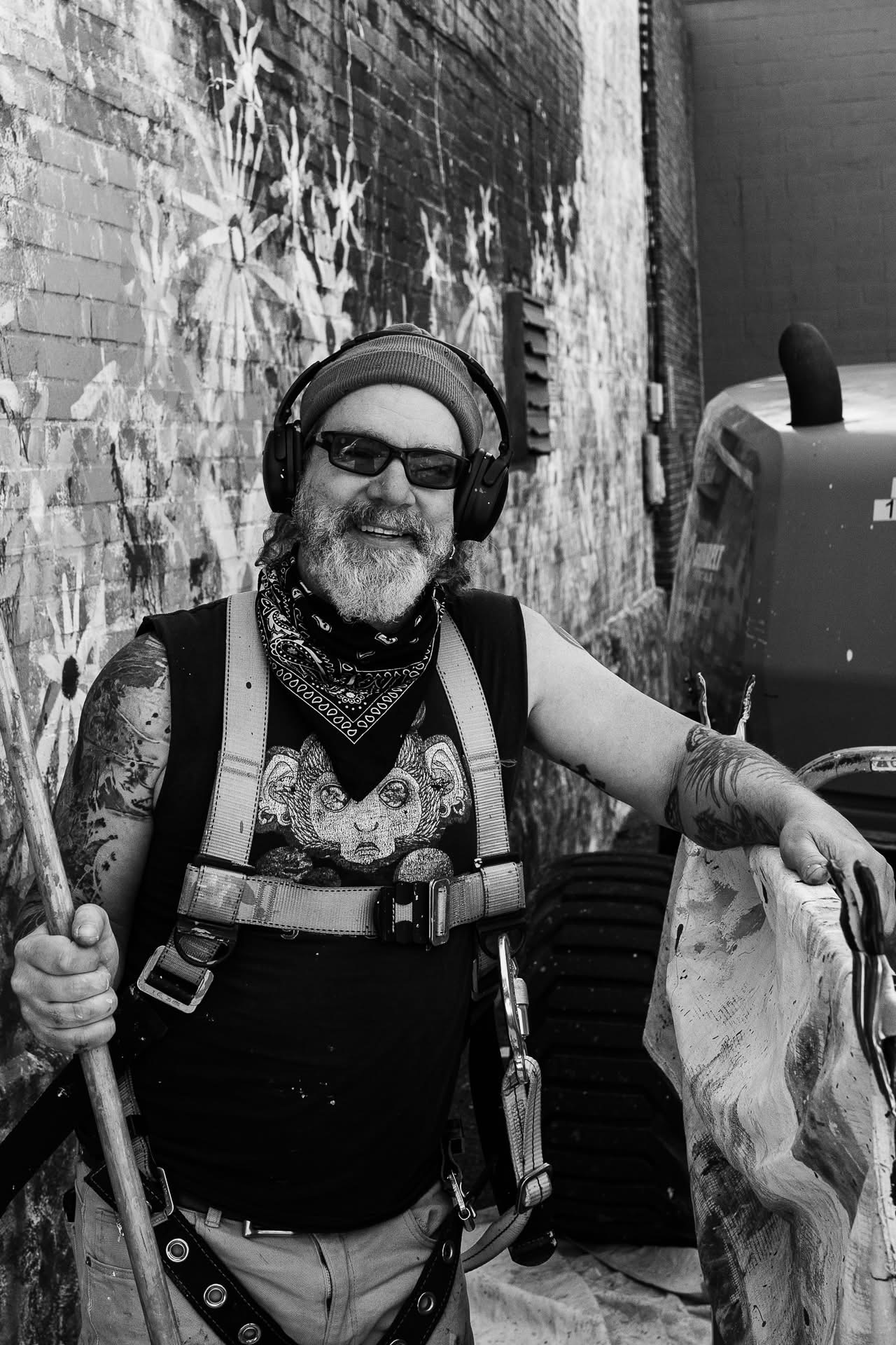 Smiling person with tattoos, wearing headphones, sunglasses, and a bandana, holding a paddle, standing outdoors near a brick wall.