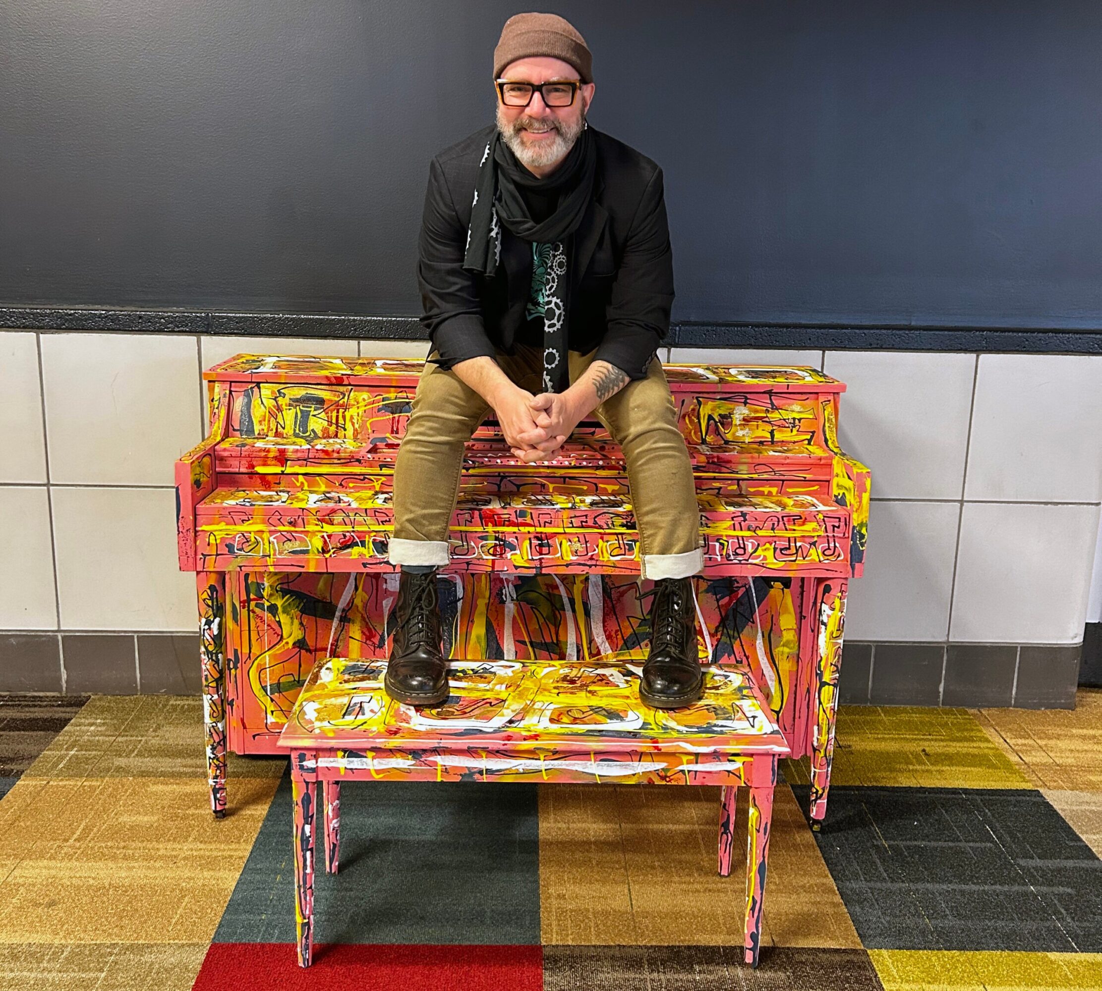 Man with glasses and a beanie sitting on a colorful, painted piano in a public space.