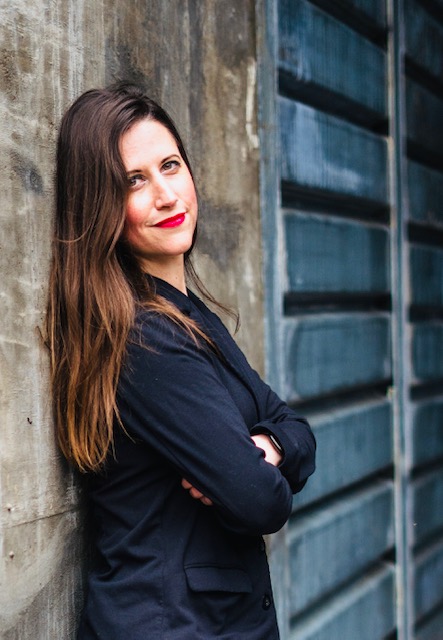 Woman with long brown hair and red lipstick leaning against a concrete wall, arms crossed, smiling slightly.