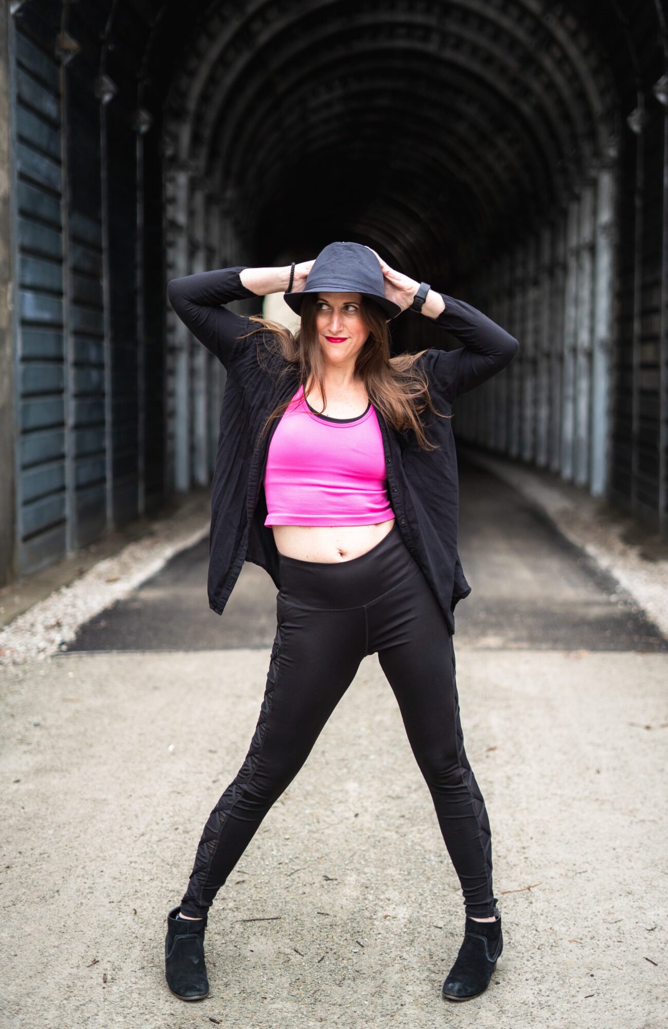 Young woman standing confidently in an urban tunnel, wearing a pink crop top, black jacket, black pants, and a hat, with hands on her hat.